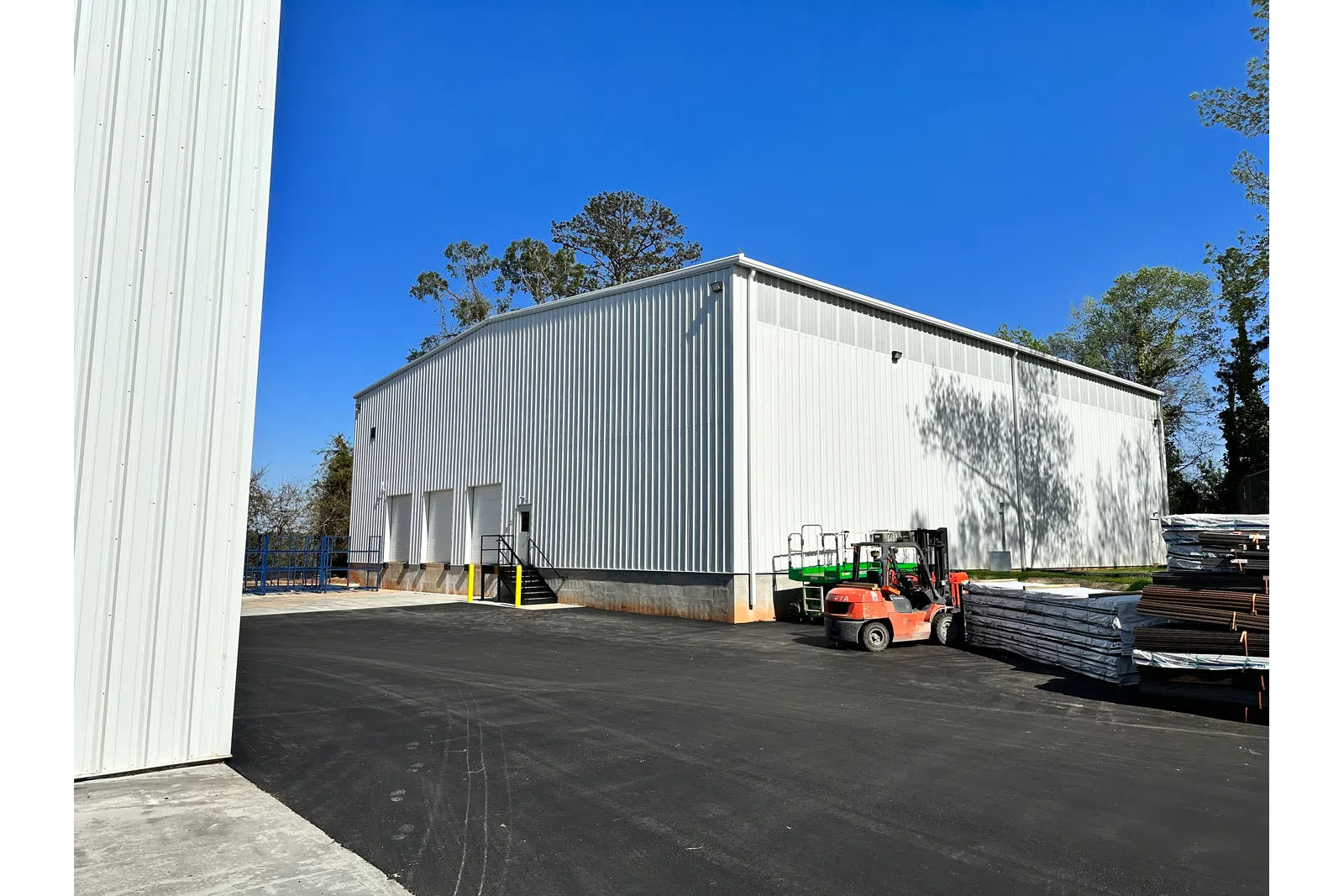 White industrial warehouse with forklifts and stacked pallets on asphalt under clear blue sky.