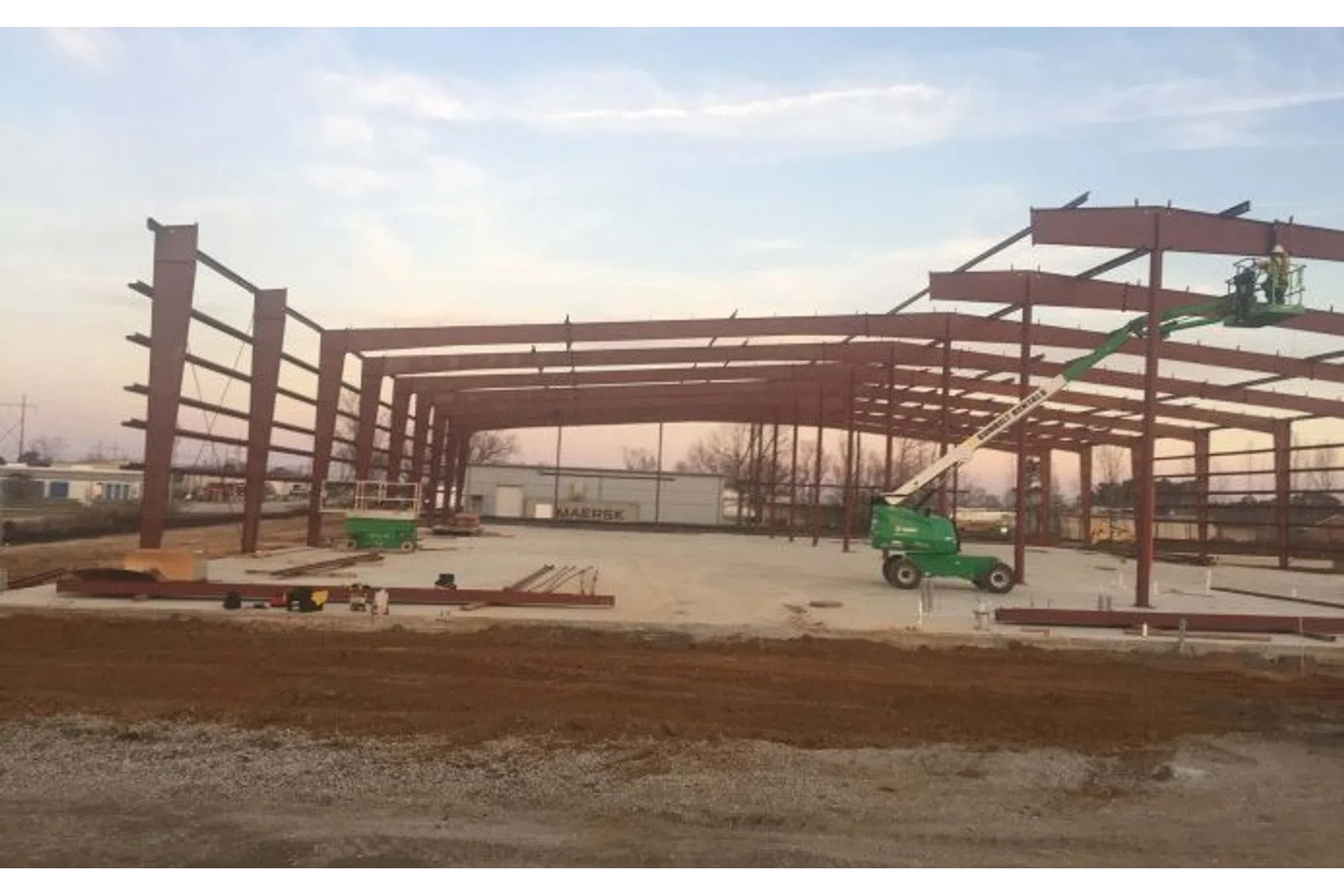Steel frame structure under construction with a green boom lift and concrete foundation at dusk.