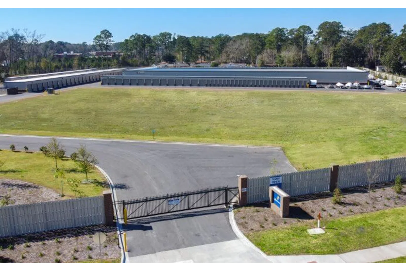 Aerial view of a gated self-storage facility with multiple storage units and large green lawn under clear sky.