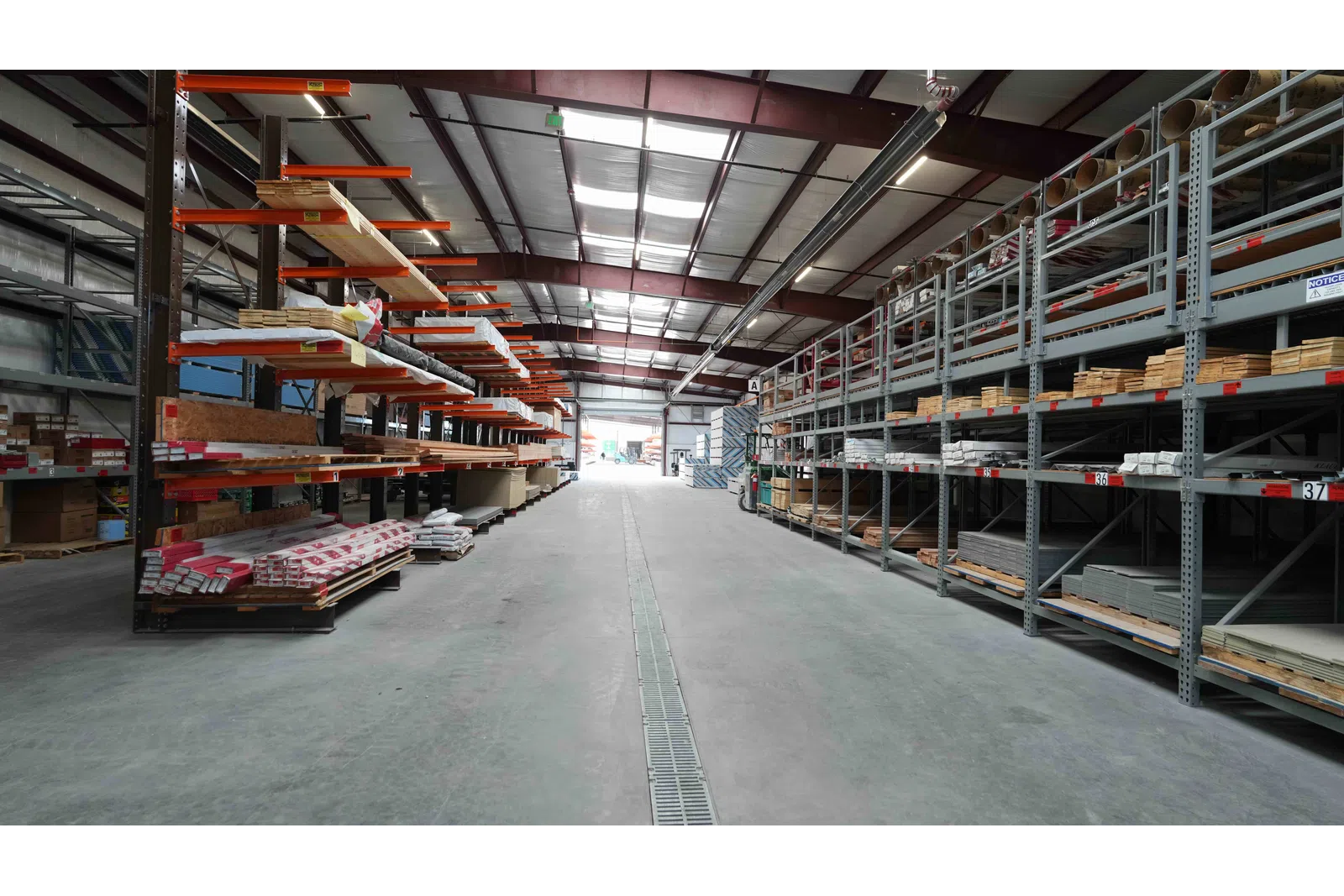 Interior view of a large warehouse with metal racks holding wood and building materials under a steel roof.
