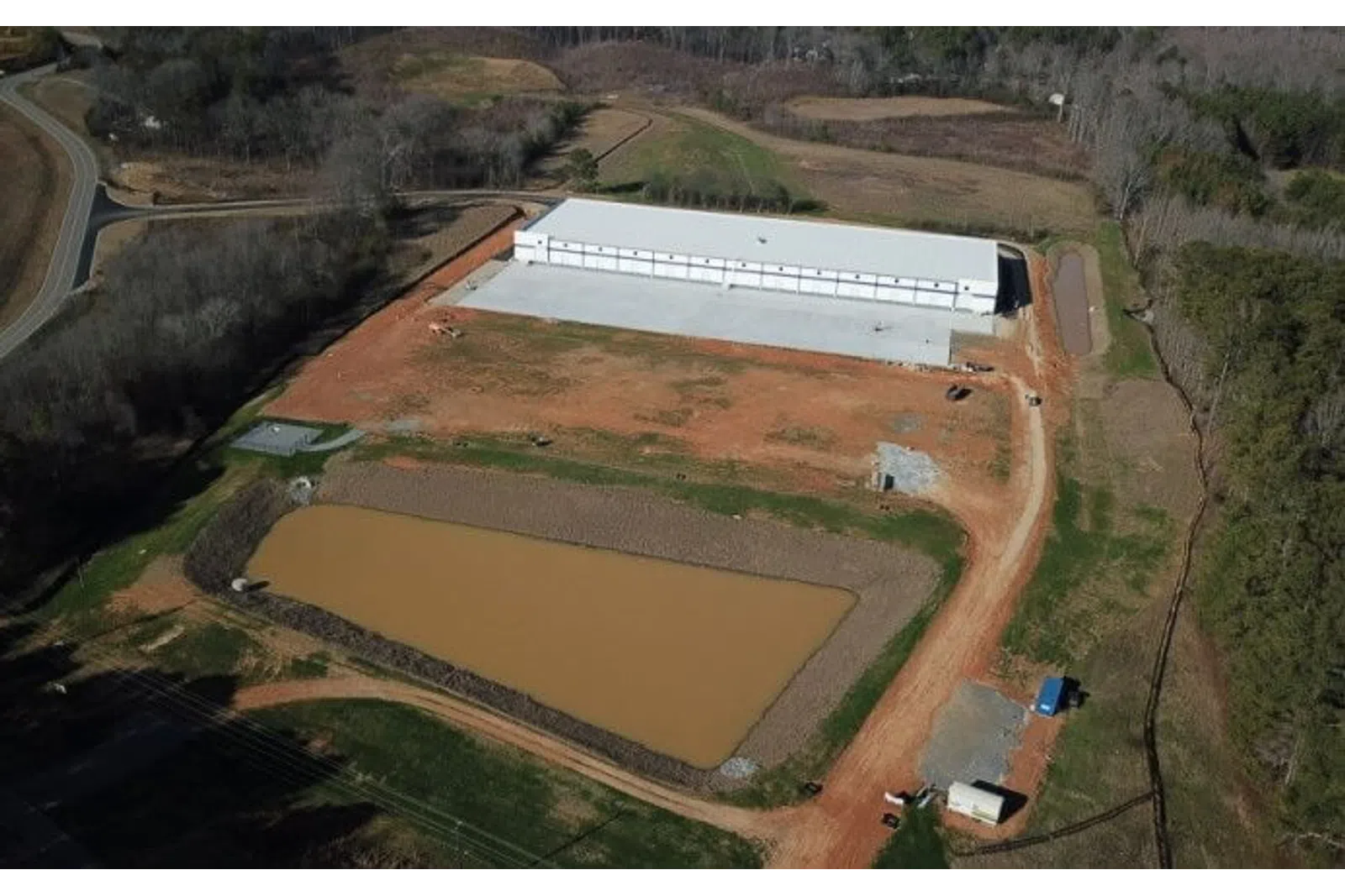 Aerial view of a large construction site with a white warehouse building near a muddy water retention pond.