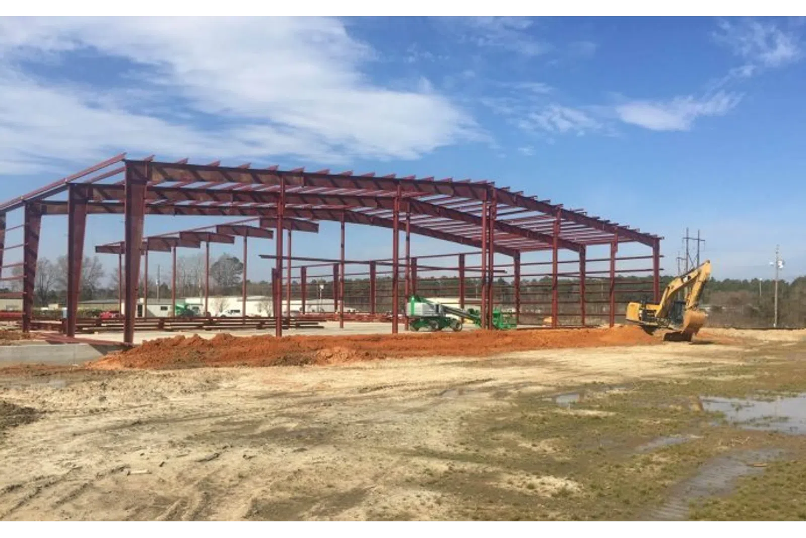 Steel frame structure under construction on a clear day with construction equipment and muddy ground in the foreground