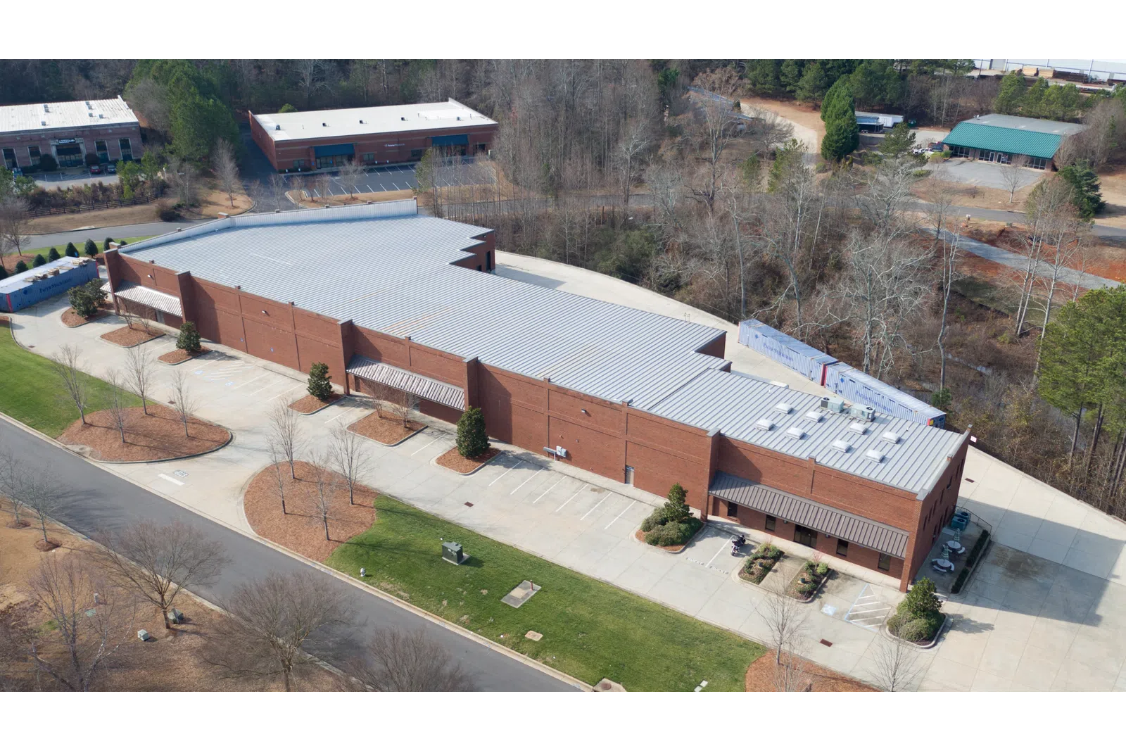 Aerial view of a large brick industrial building with a metal roof surrounded by parking areas and trees.