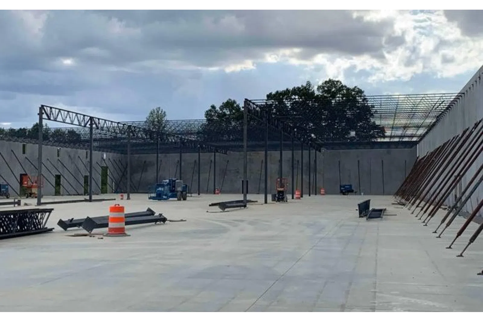 Construction site with concrete floor, steel framework, and orange traffic barrels under a cloudy sky.