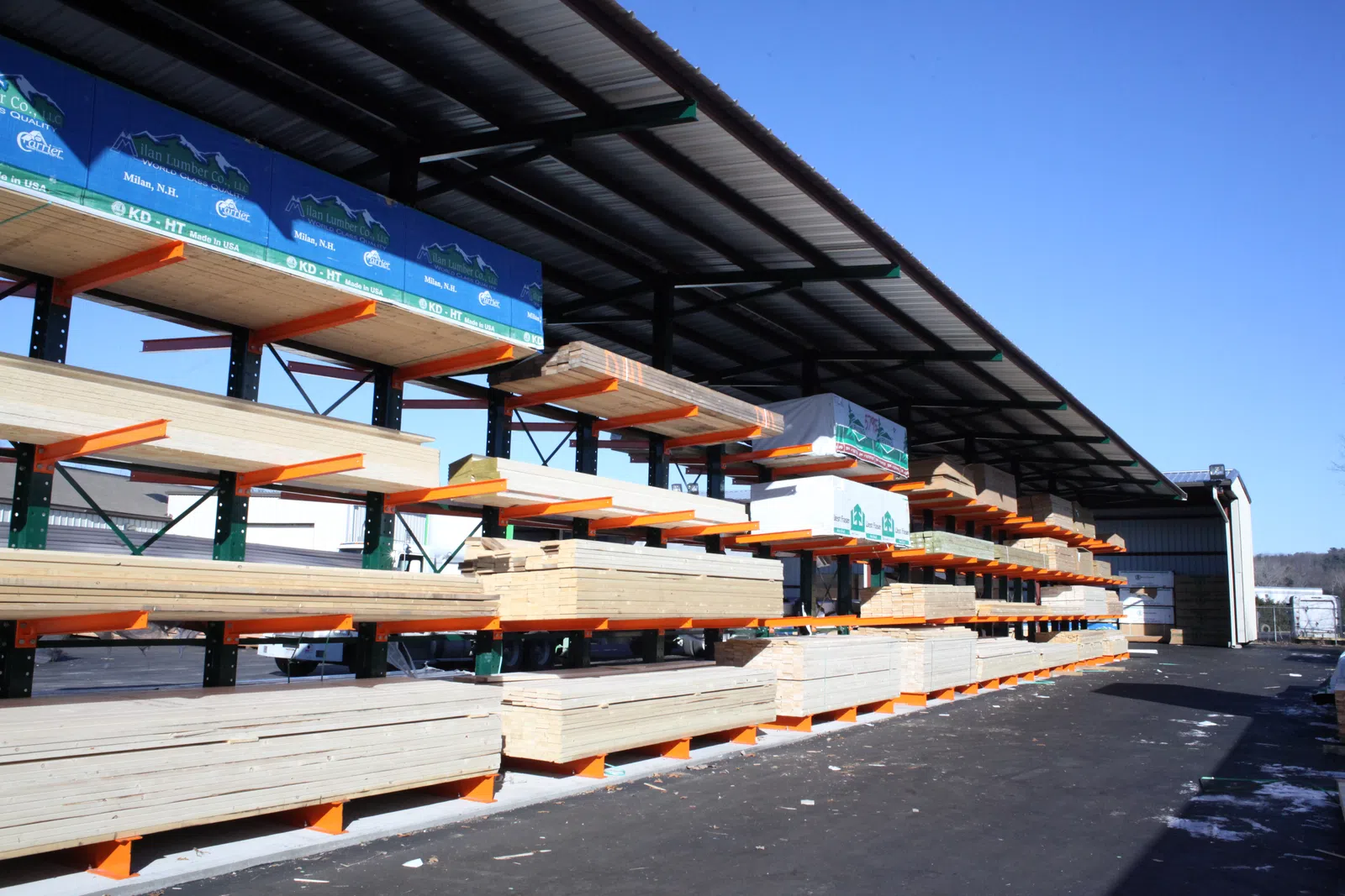 Outdoor covered lumber rack holding stacked sheets of plywood and wood boards at a hardware store under clear blue sky
