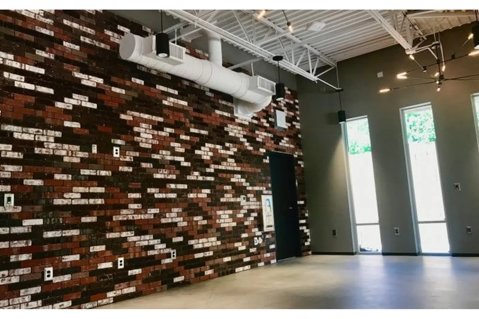 Industrial-style room with exposed brick wall, high ceiling, large windows, and concrete floor.