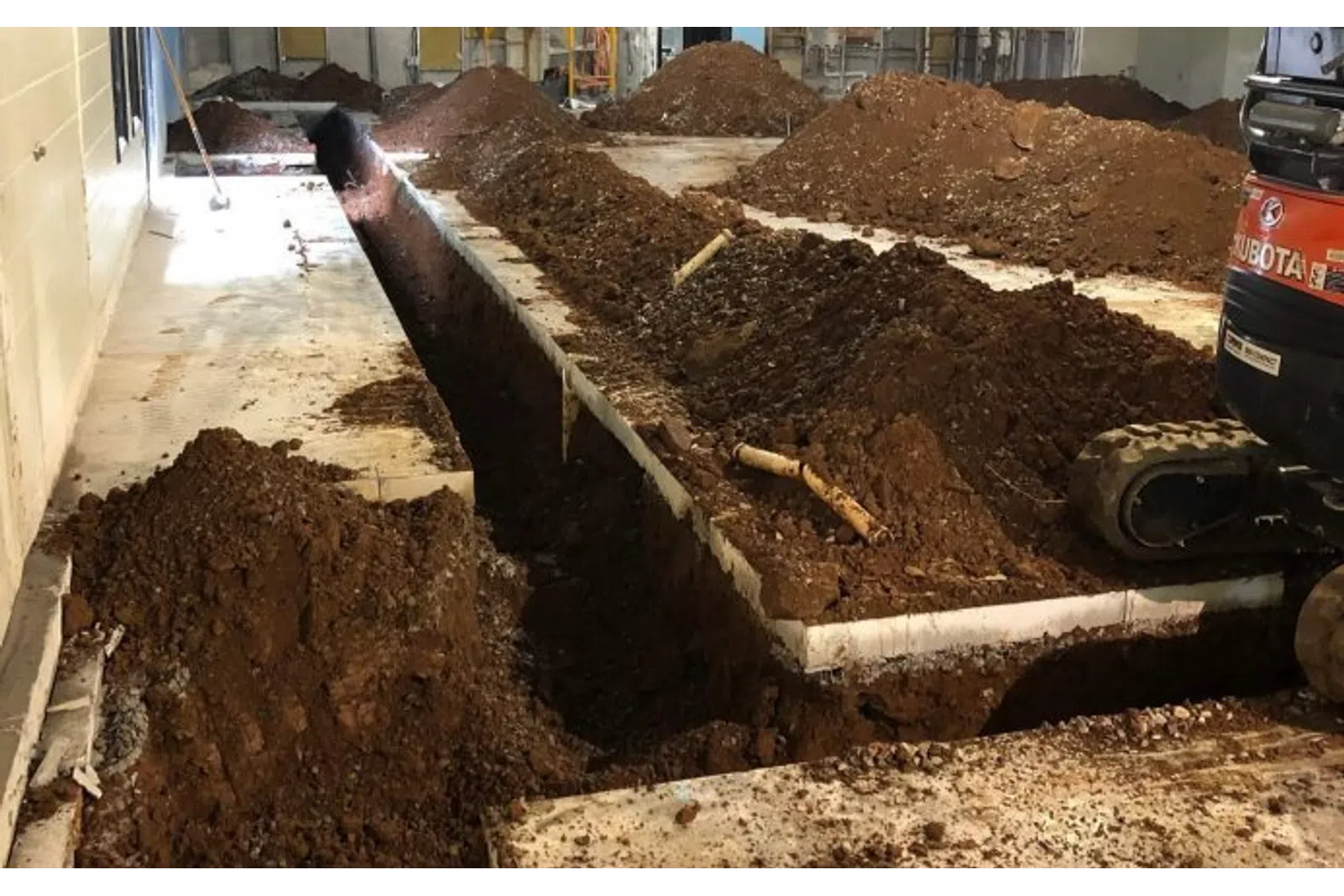 Indoor construction site with deep trenches dug in concrete floor and piles of soil around machinery.