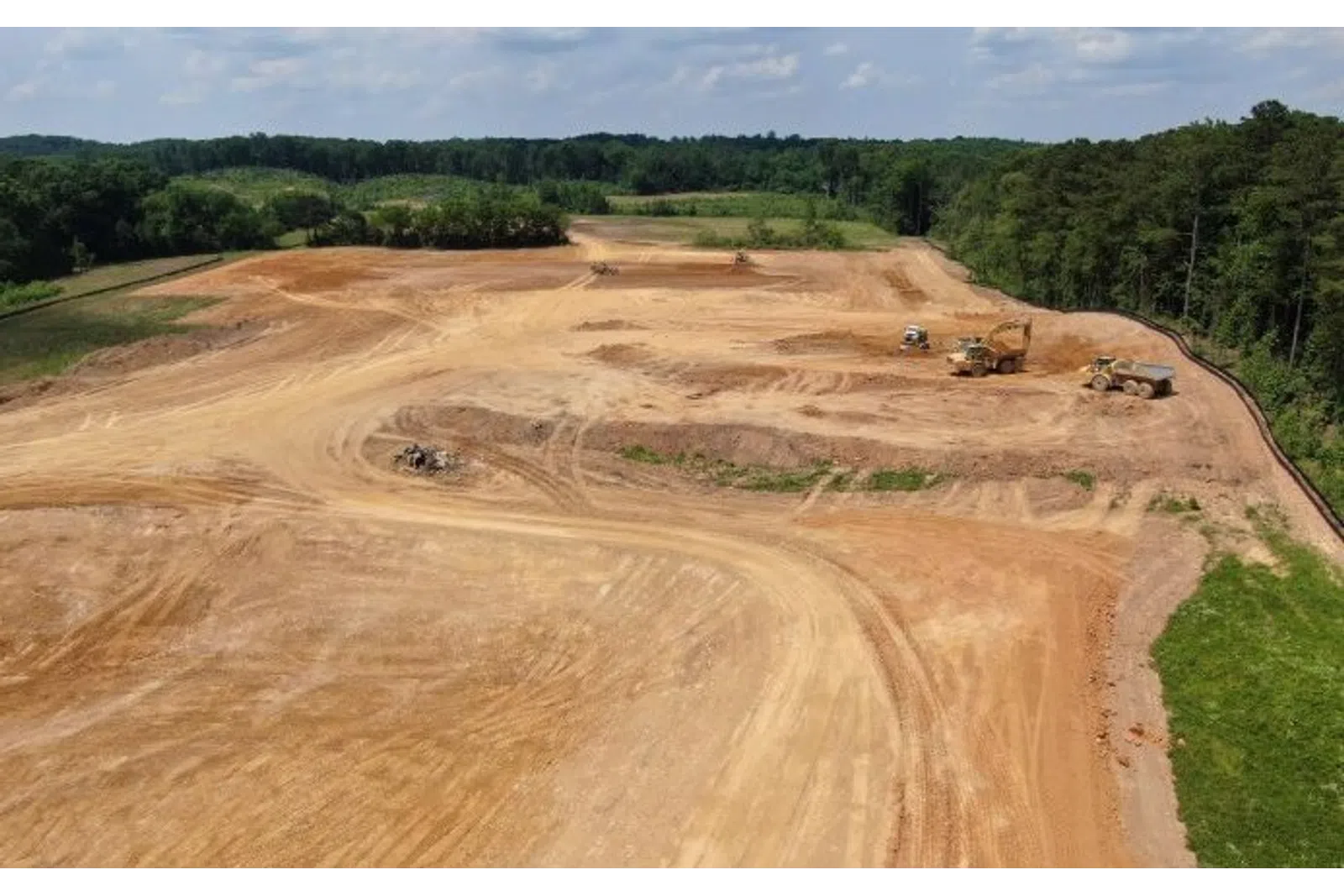 Large construction site with dirt roads, heavy machinery, and forested background under partly cloudy sky