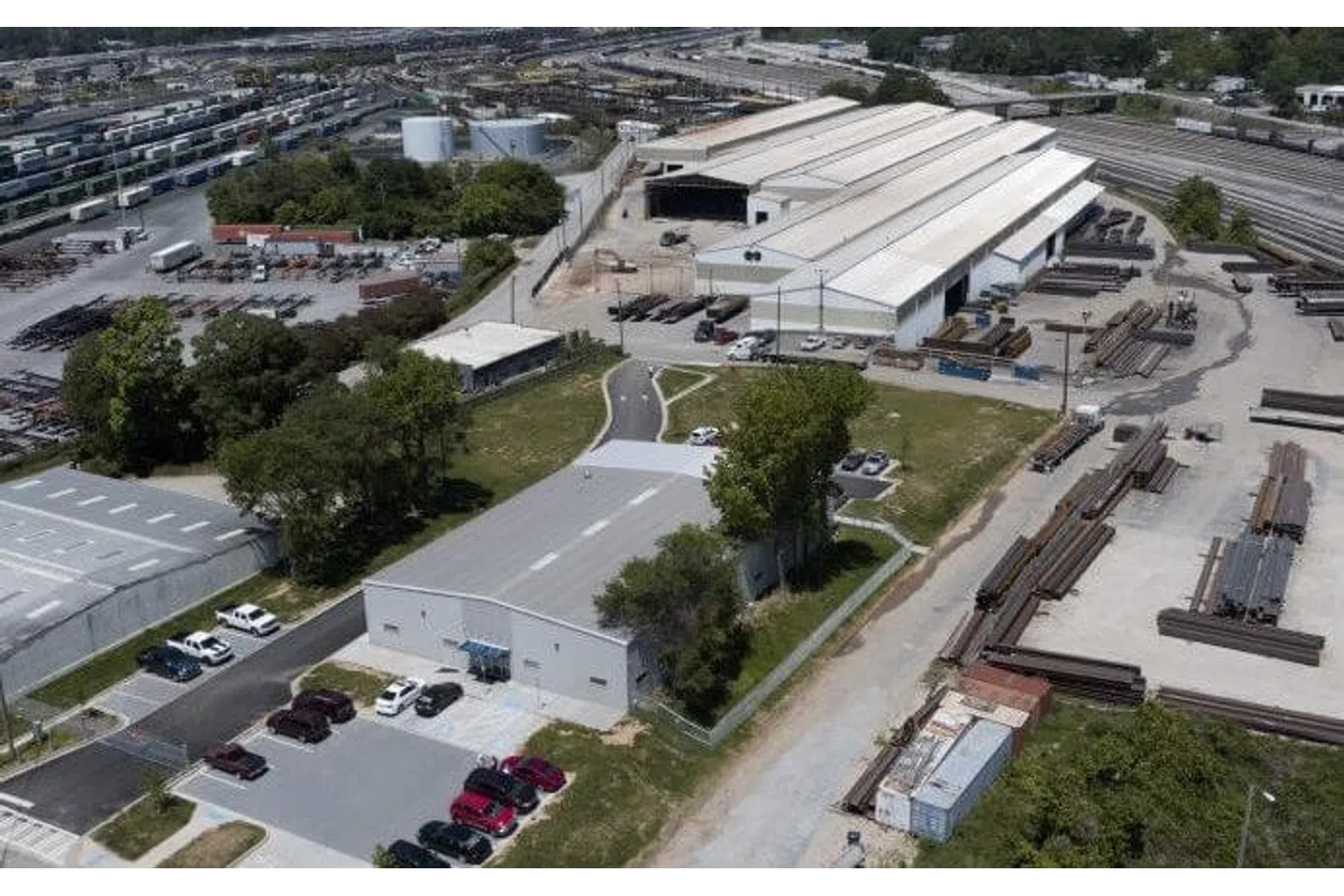 Aerial view of industrial warehouses with parking lot, storage yard, railroad tracks, and surrounding greenery on a sunny day.