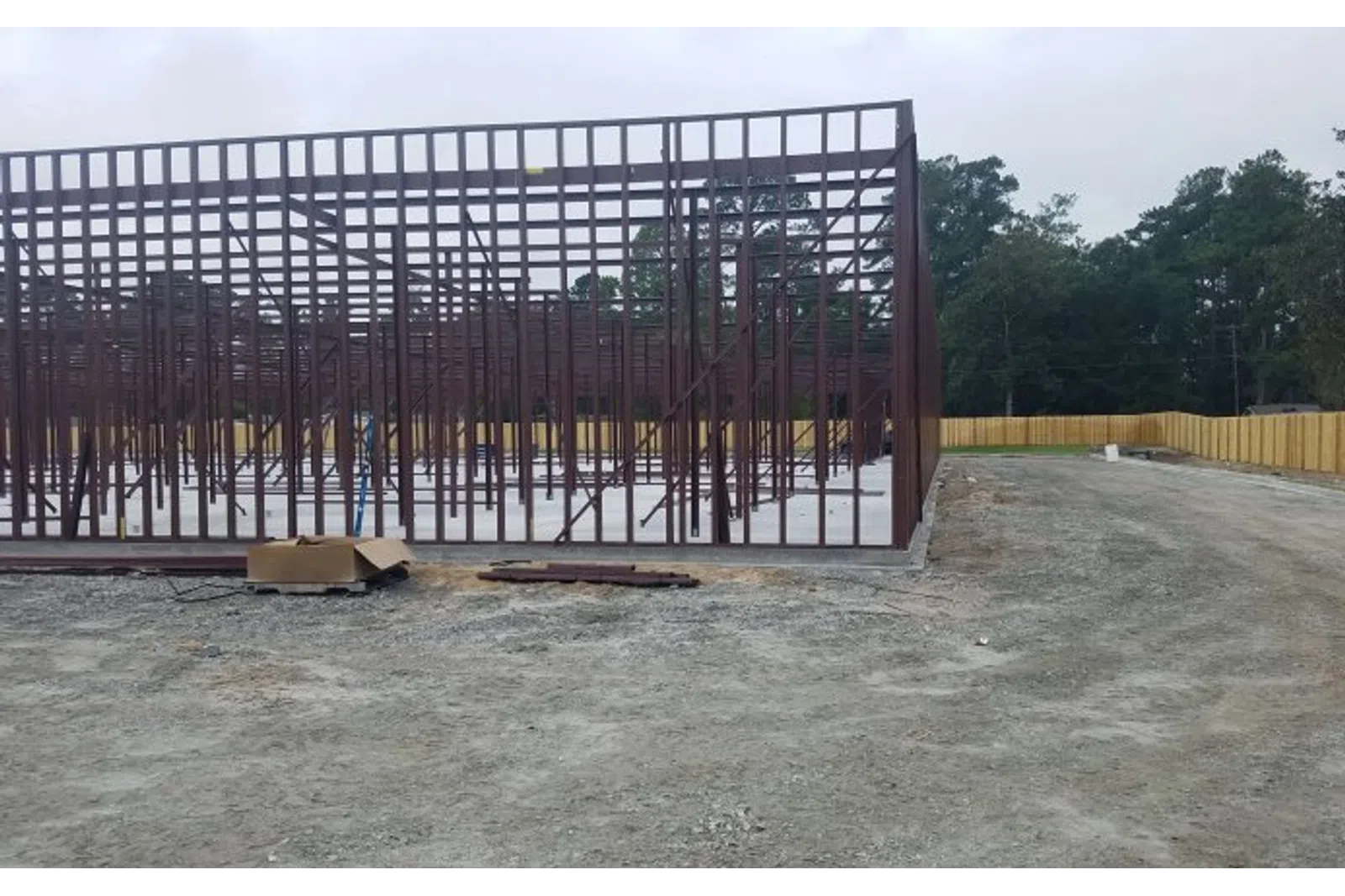 Steel framework of a building under construction on a cleared dirt site with a wooden fence and trees in the background.