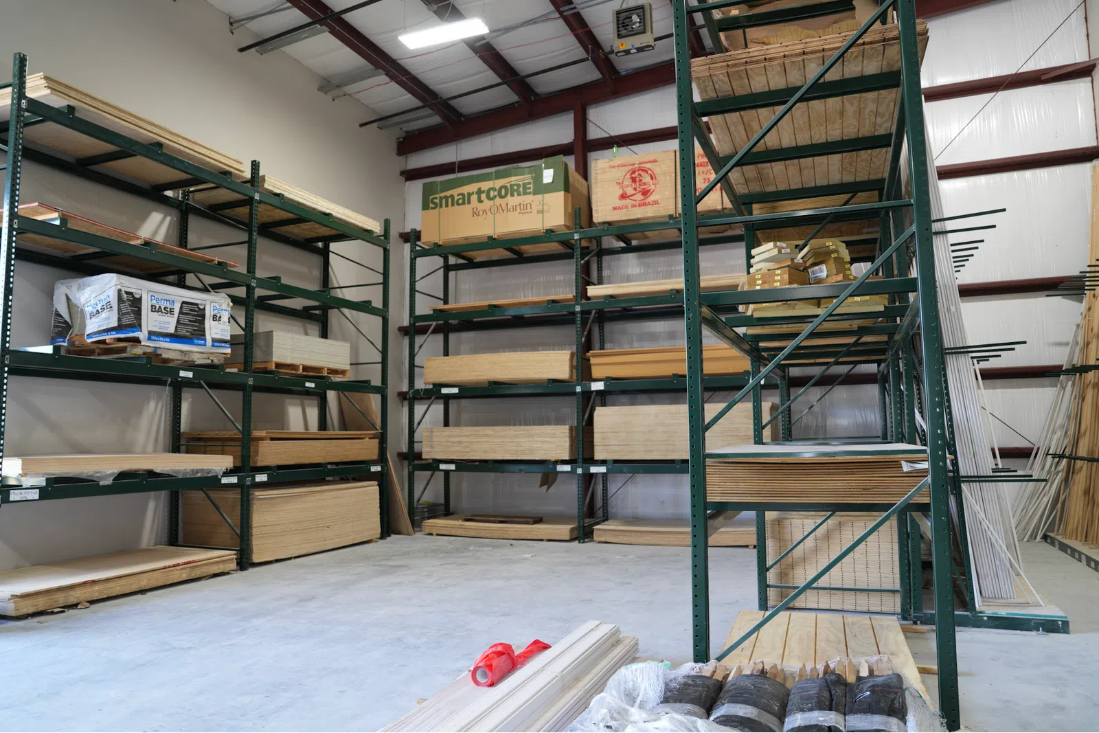 Wood sheets and building materials organized on green metal shelves in a spacious warehouse with concrete floor.