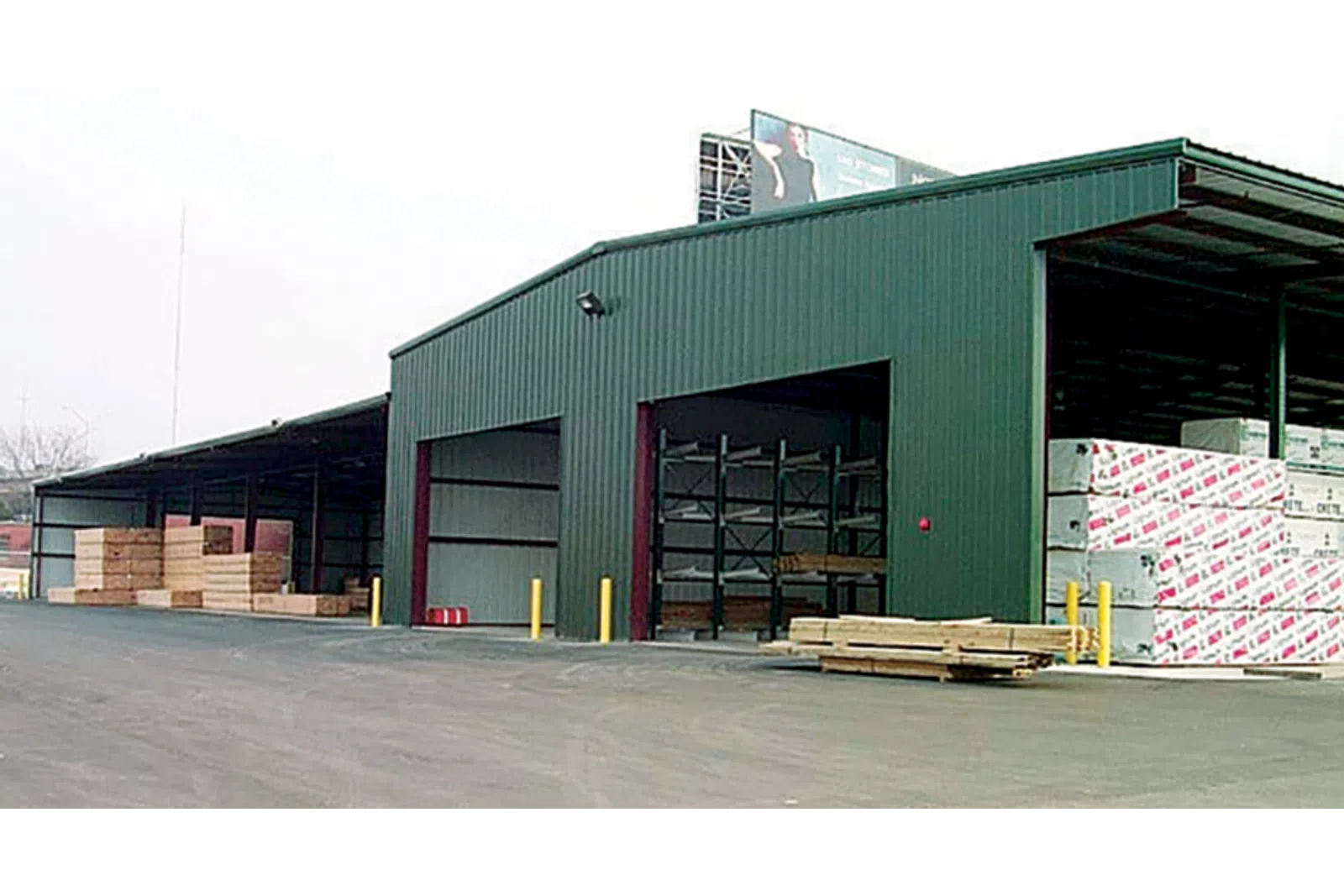 Large green warehouse storing various wood planks and construction materials with open bays.