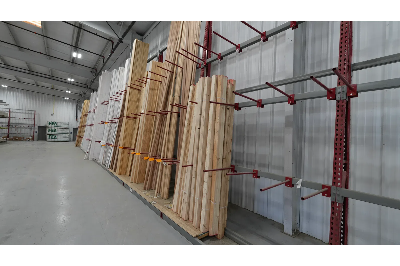 Stacks of wooden planks stored vertically on red metal racks inside a warehouse with high ceilings.