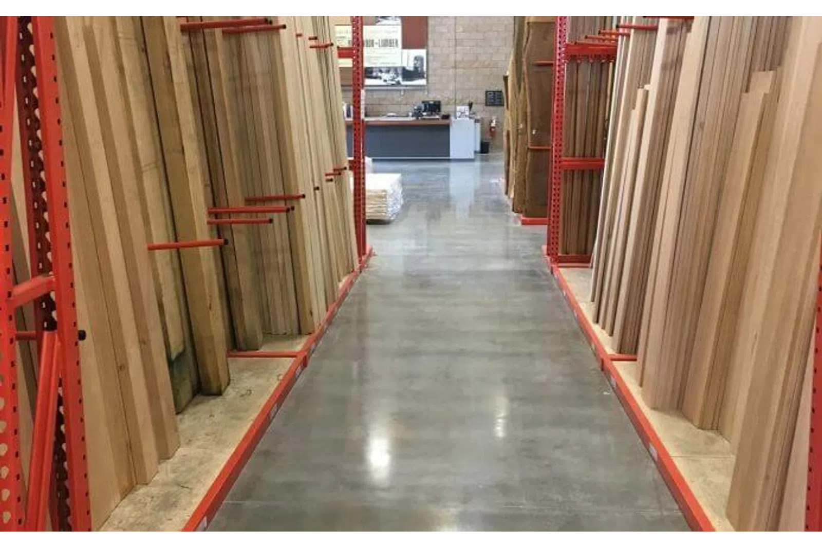 Aisle in a hardware store with stacks of wooden boards organized on red metal racks on both sides.