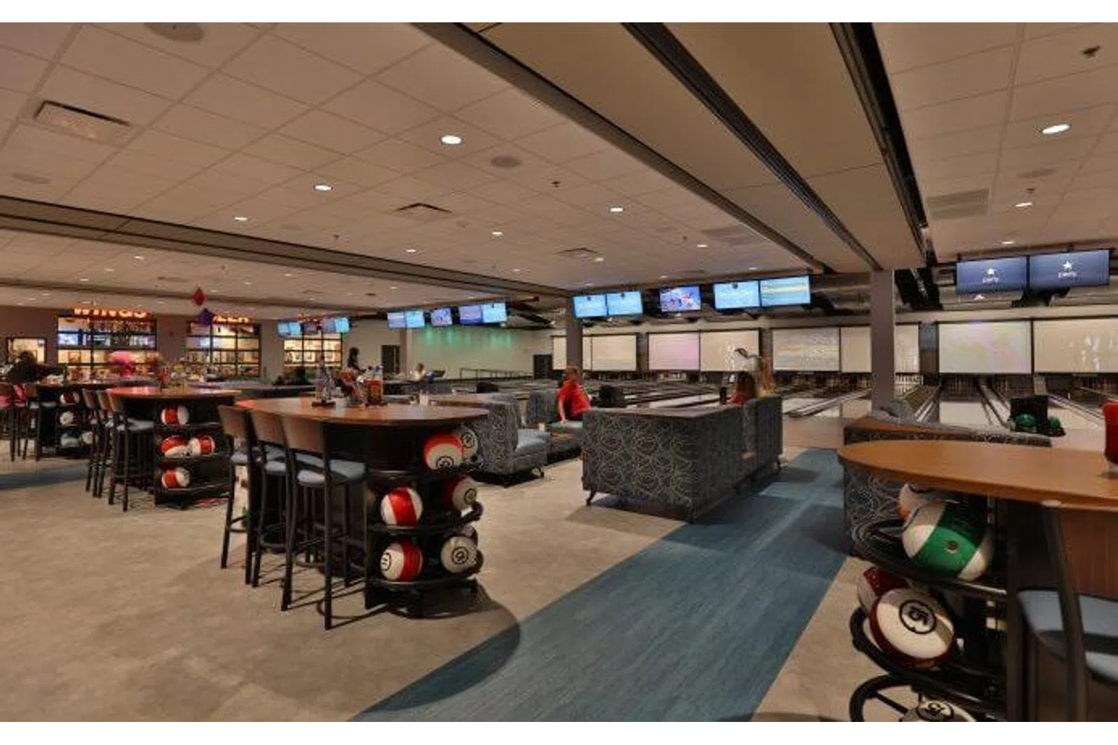 Modern bowling alley interior with seating, tables, bowling balls, and multiple lanes with screens.