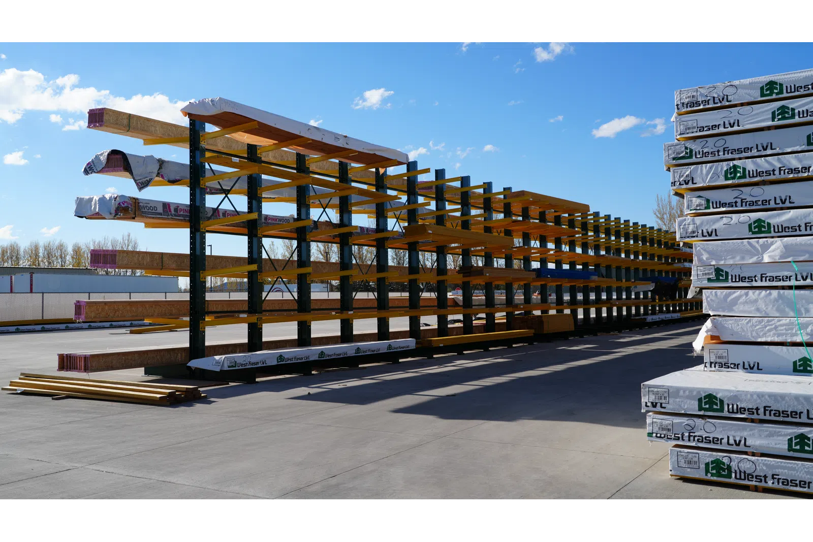 Outdoor lumber storage rack with long wooden beams and stacked West Fraser LVL packages on a sunny day.
