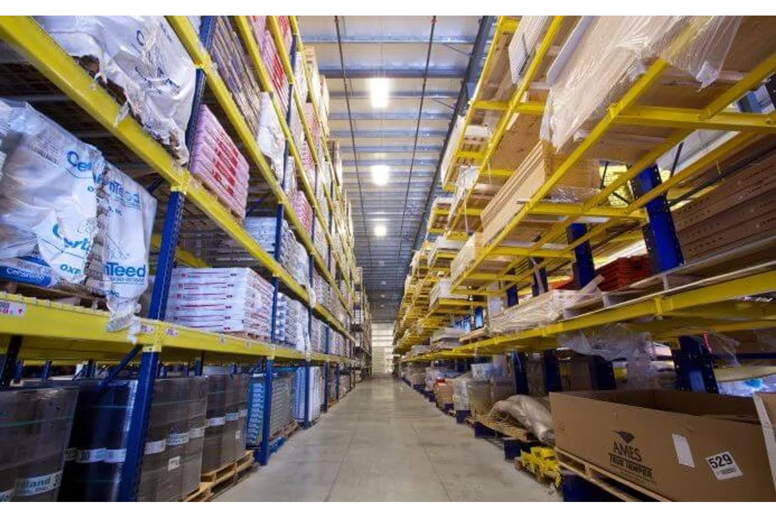 Wide aisle in warehouse lined with tall yellow shelves stocked with boxes and packaged goods.