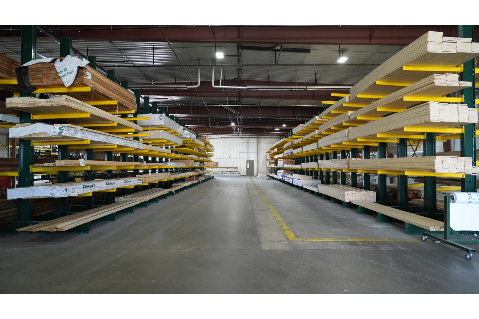 Interior view of a warehouse aisle with stacked lumber on metal racks and a concrete floor under overhead lighting.