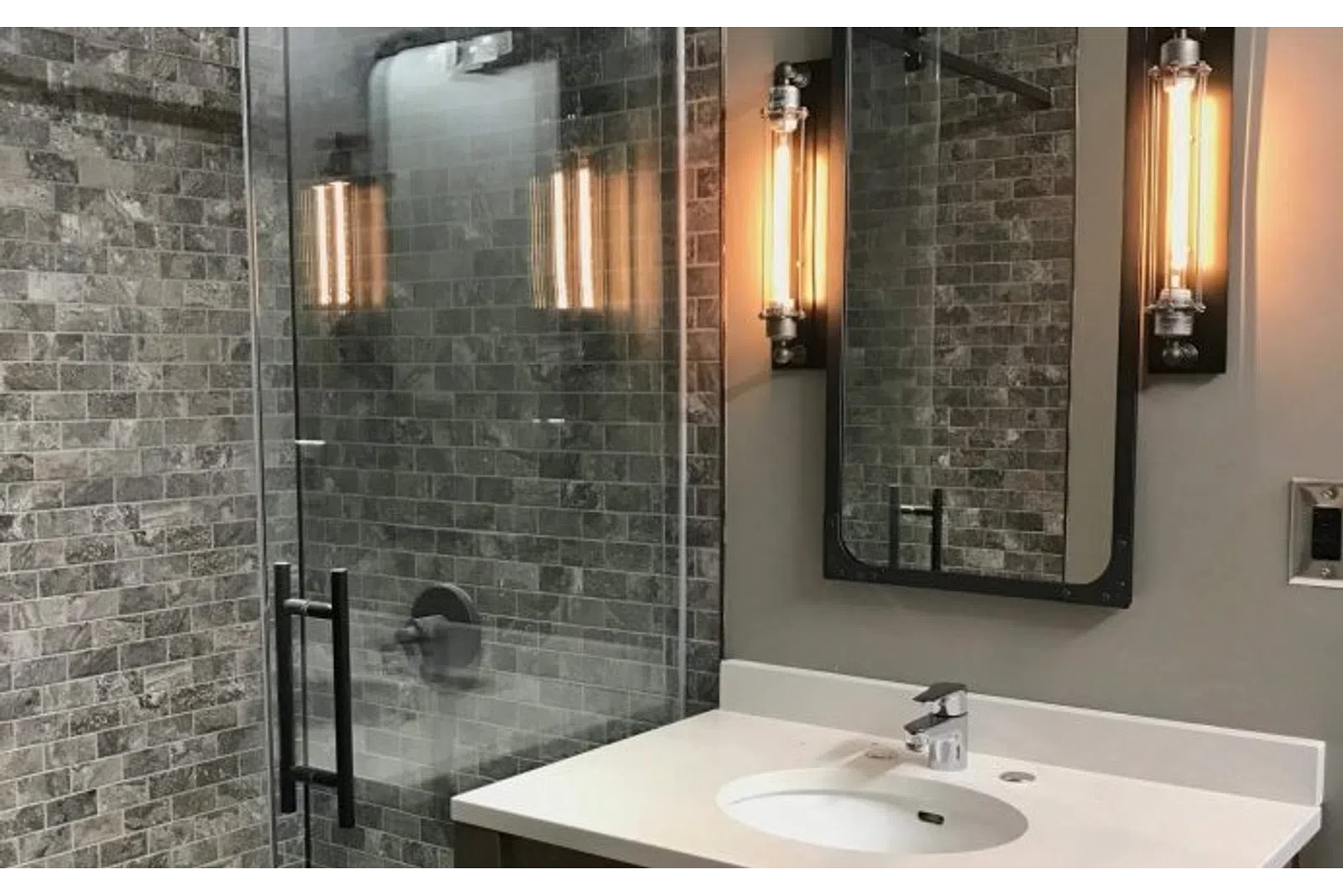 Modern bathroom with glass shower, gray stone tiles, white countertop, and industrial wall light fixtures.