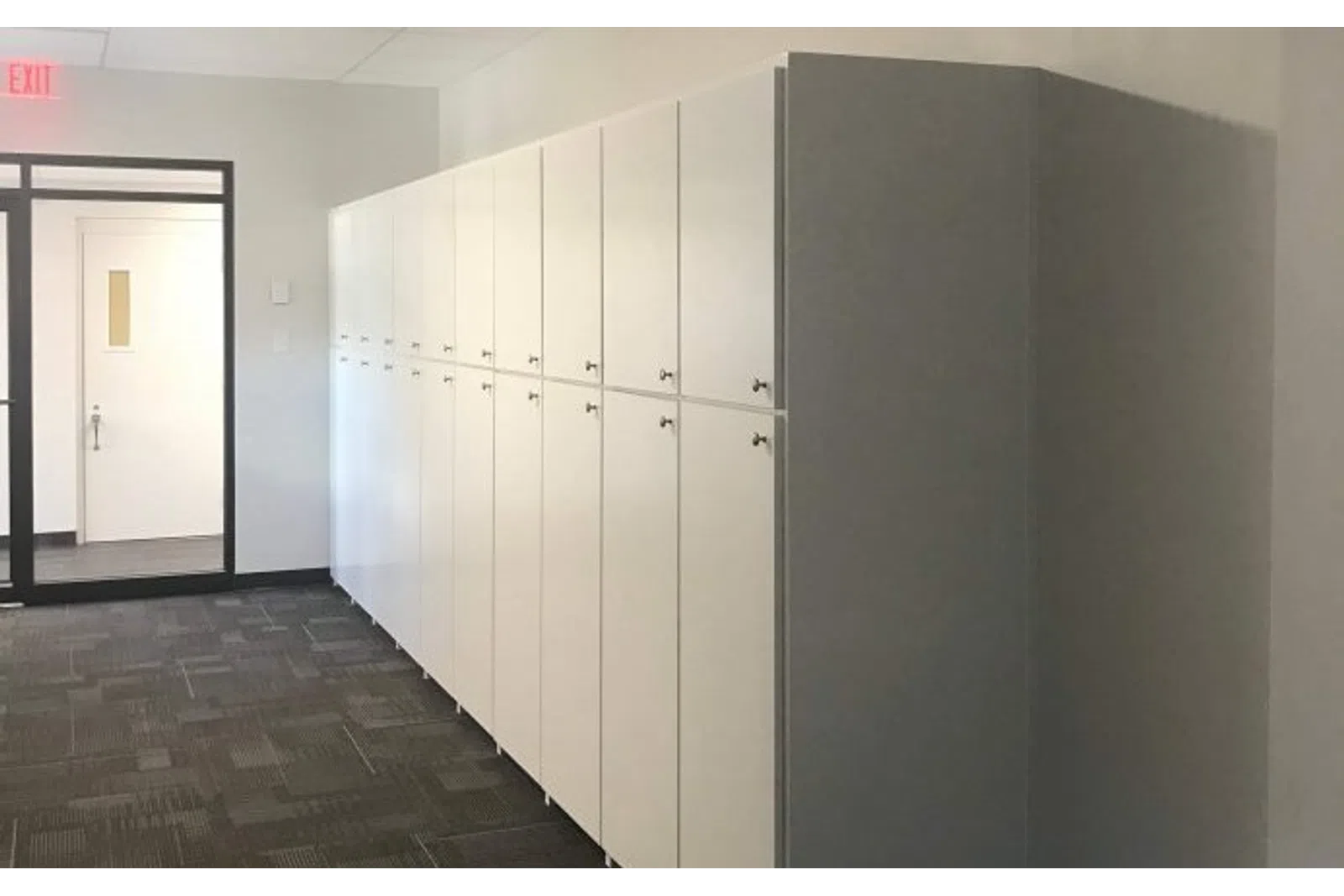 Row of white storage lockers with locks in a modern office corridor with carpet and glass exit door.