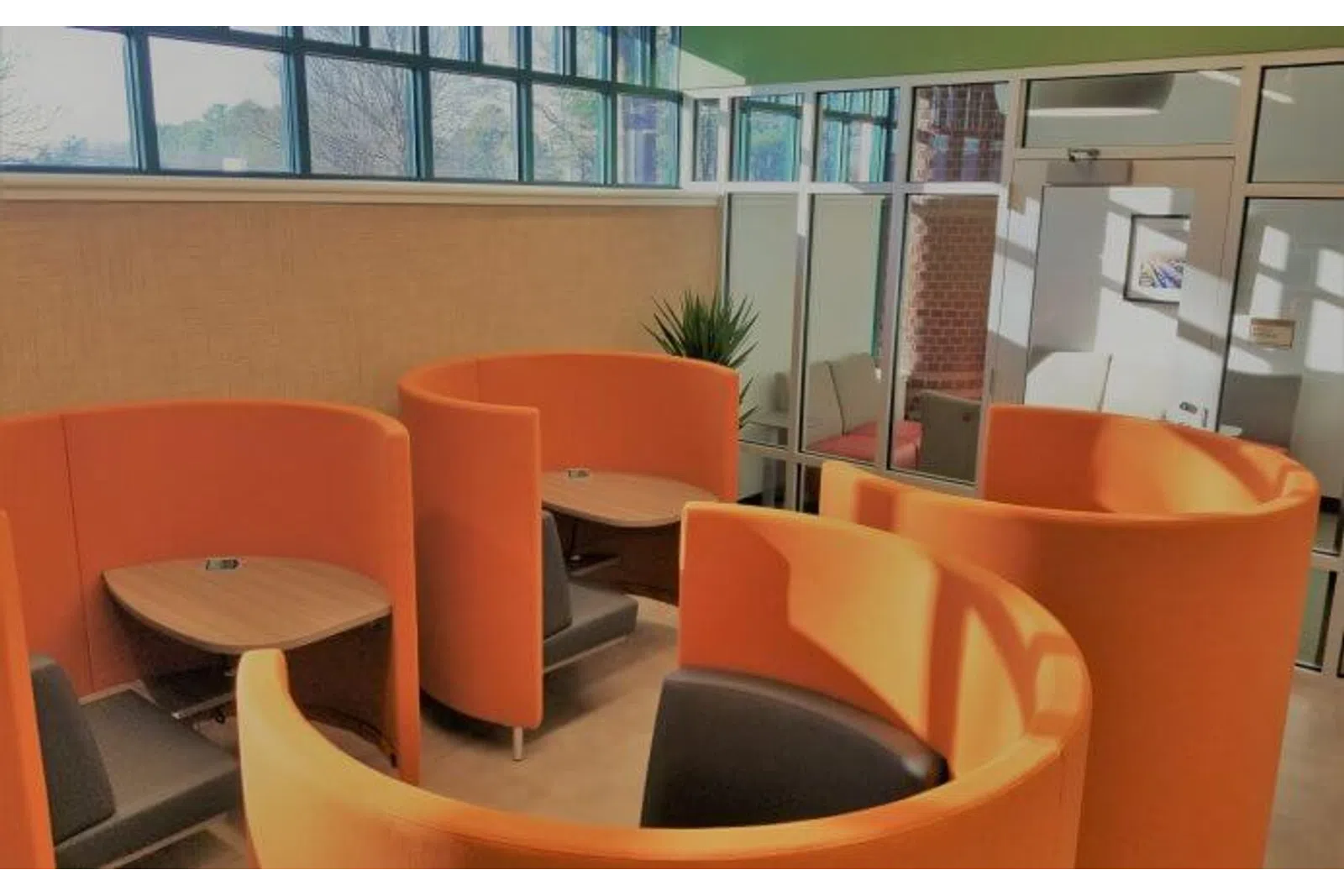 Modern office lounge area with curved orange seating pods, small wooden tables, and black chairs under natural light.