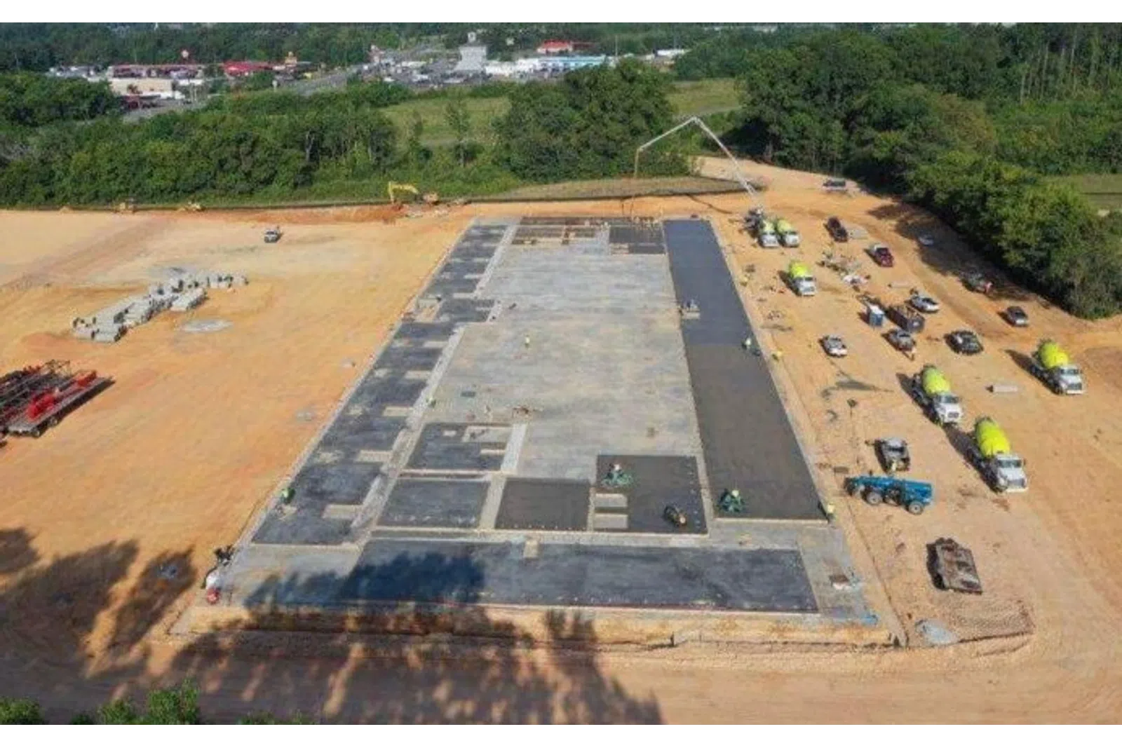 Aerial view of concrete foundation construction with trucks and workers on a cleared dirt site near trees.