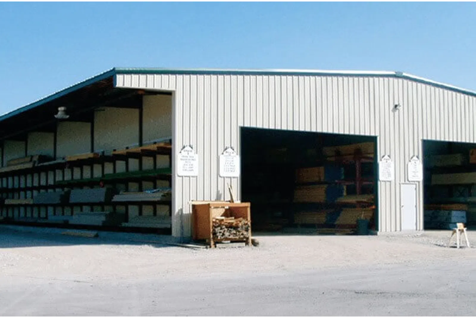 Large metal warehouse with open bays storing lumber and wood products under clear blue sky.