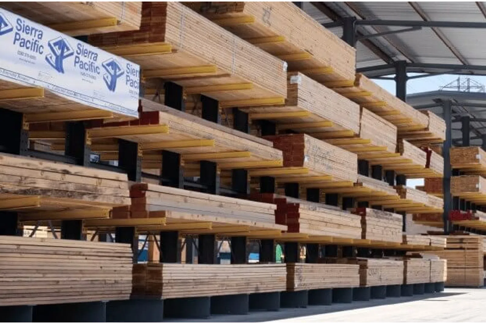Stacks of lumber stored on metal racks at an outdoor lumber yard under a metal roof structure.