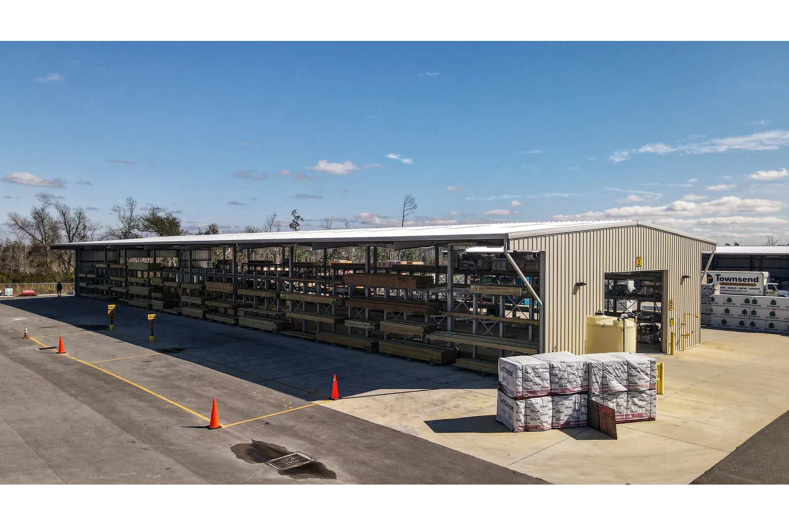 Outdoor lumber storage rack under metal shelter with pallets and an industrial warehouse in a sunny setting.