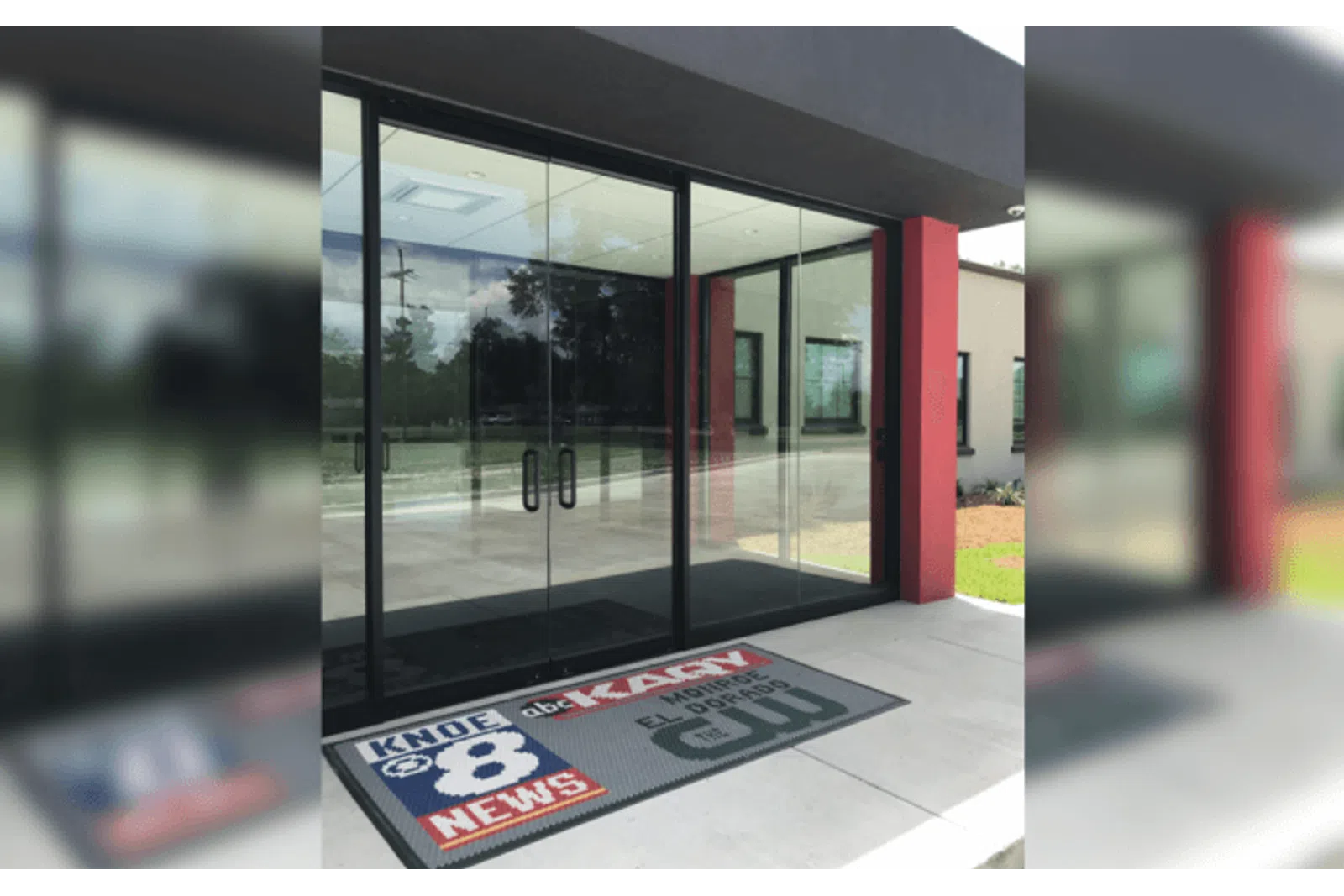 Glass entrance doors with KNOE 8 News and local TV station logos on the floor mat outside a building.