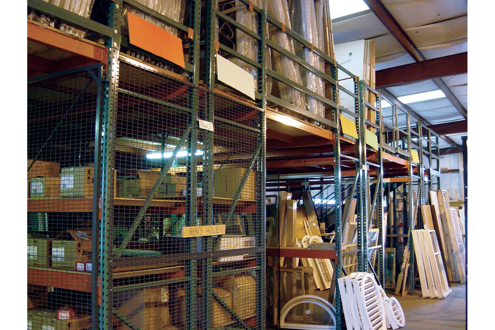 Warehouse storage with metal shelving holding boxes and wooden panels under industrial roof lighting.