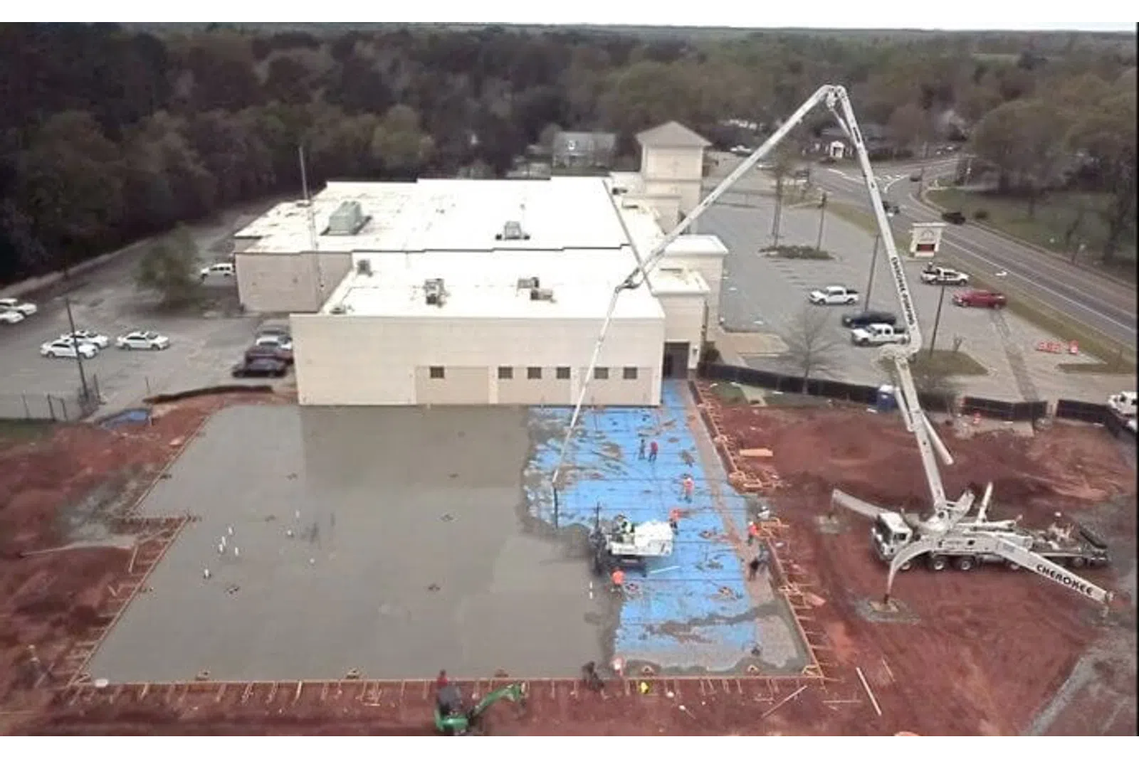 Aerial view of concrete pouring at construction site next to a white building and parking lot with trees in the background