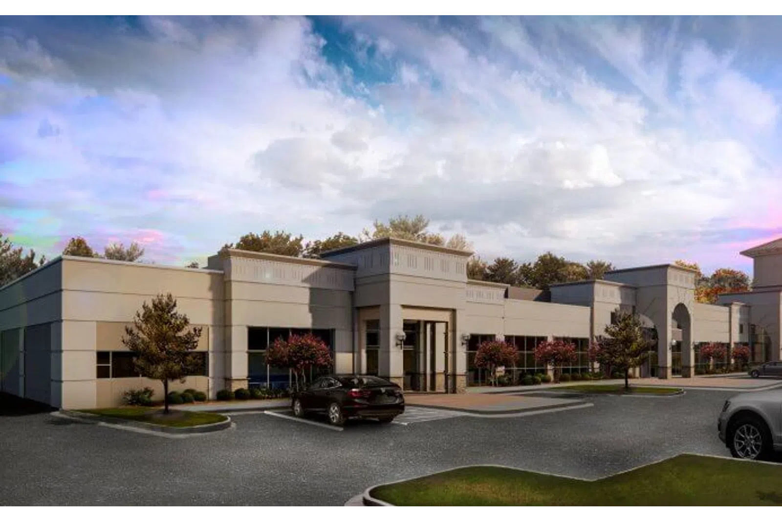 Modern commercial office building with beige facade, parking lot, trees, and cloudy sky at dusk.