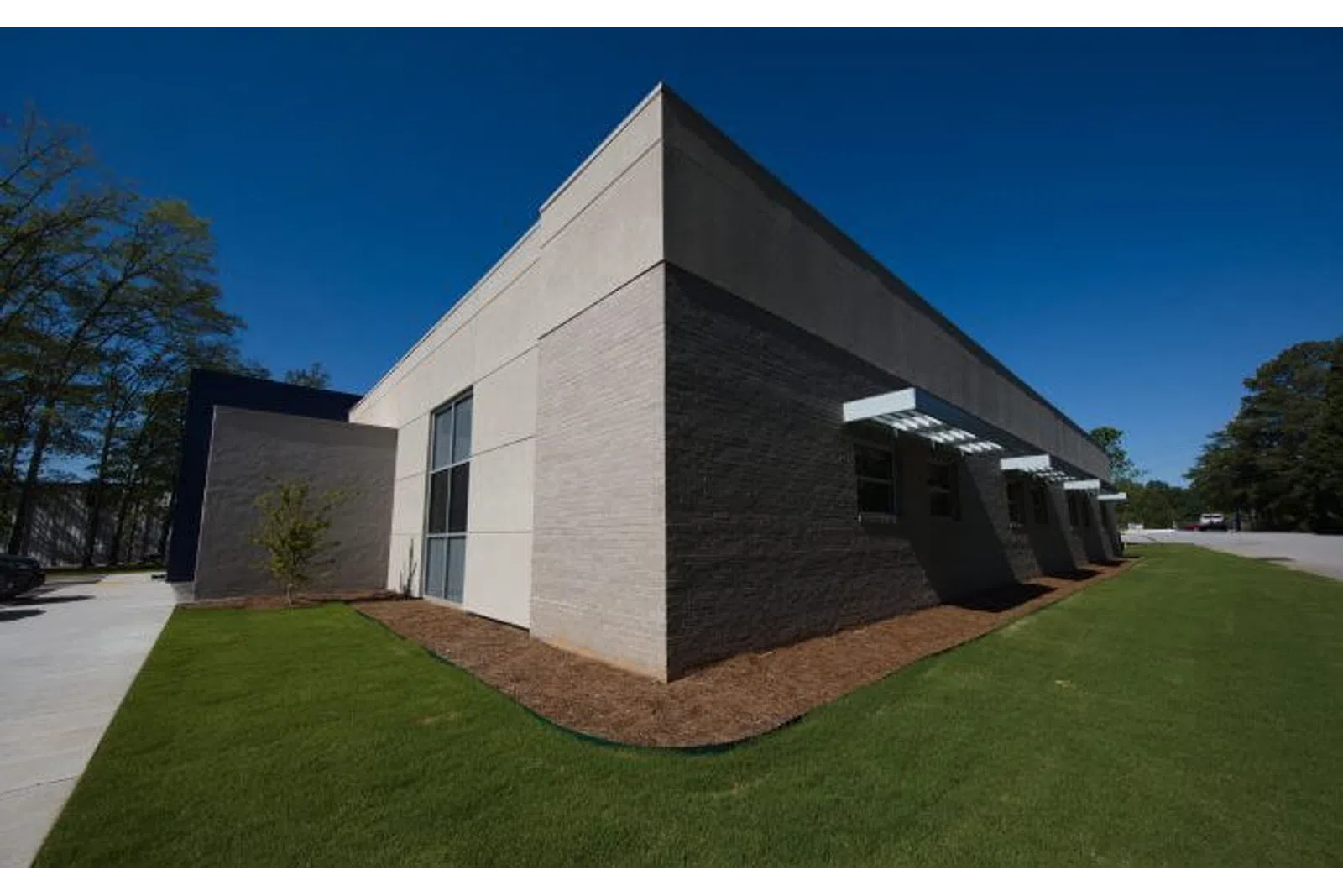 Modern single-story commercial building with gray brick walls, large windows, and green lawn under a clear blue sky.