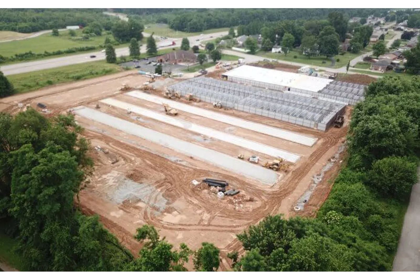 Aerial view of a construction site with foundation slabs and steel framework surrounded by greenery and nearby roads.