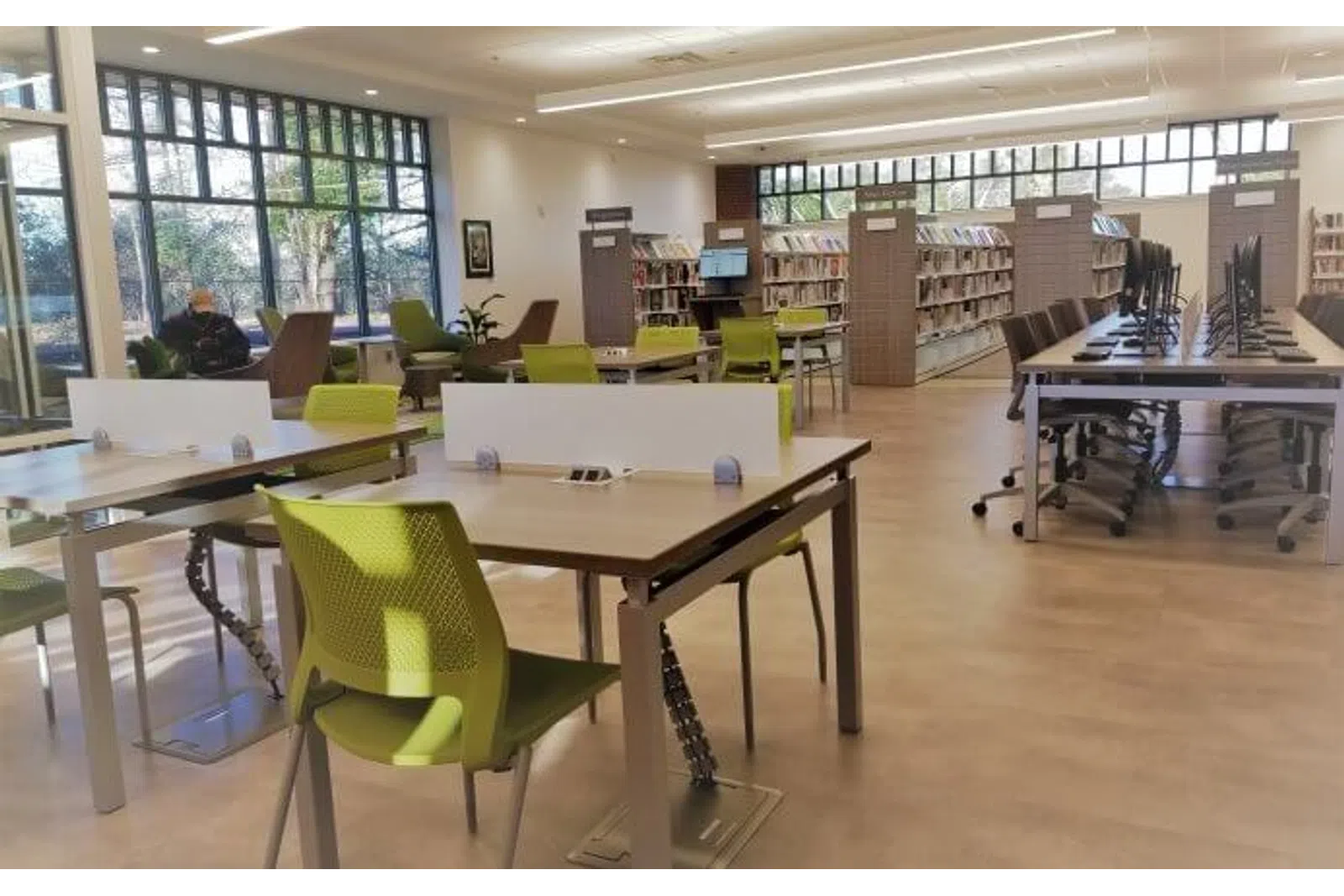 Modern library interior with green chairs, study tables, computer stations, and large windows letting in natural light.