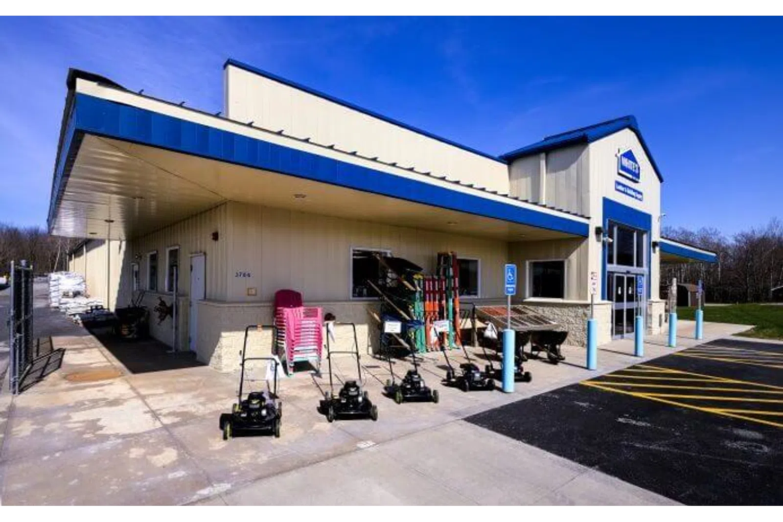 Outdoor picture of a hardware store with lawnmowers and garden tools displayed on the sidewalk under a blue awning.