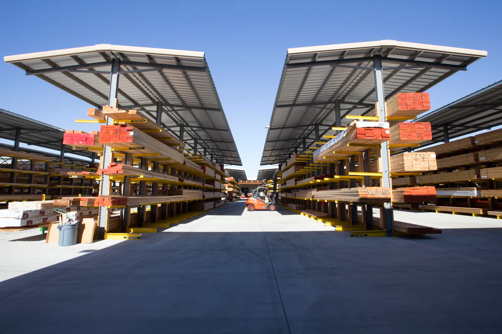 Outdoor lumber yard with organized stacks of wooden planks under metal roofs and a forklift in the distance.