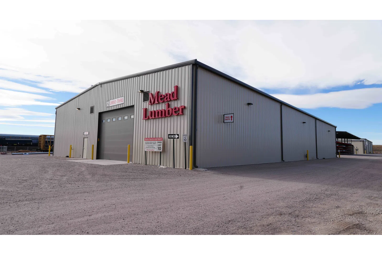 Large gray metal industrial building with Mead Lumber signage and drive-thru entrance under a cloudy sky