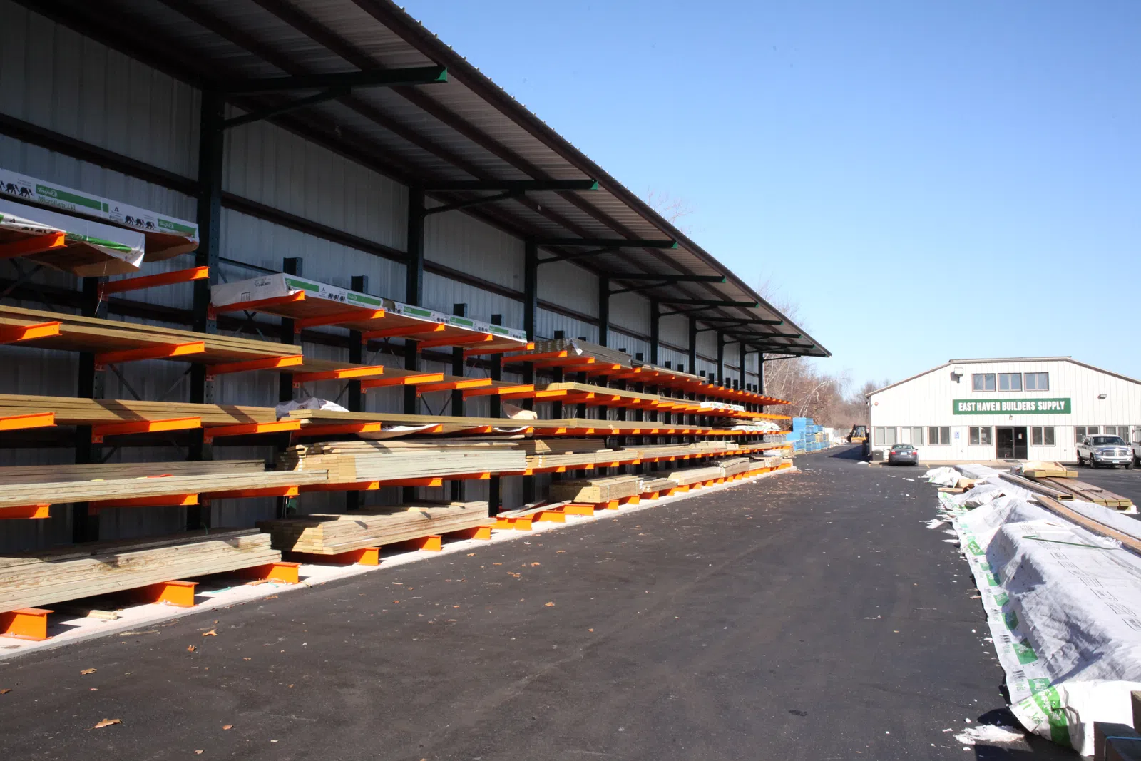 Outdoor lumber yard with stacked wood planks on orange racks and a building labeled East Haven Builders Supply.