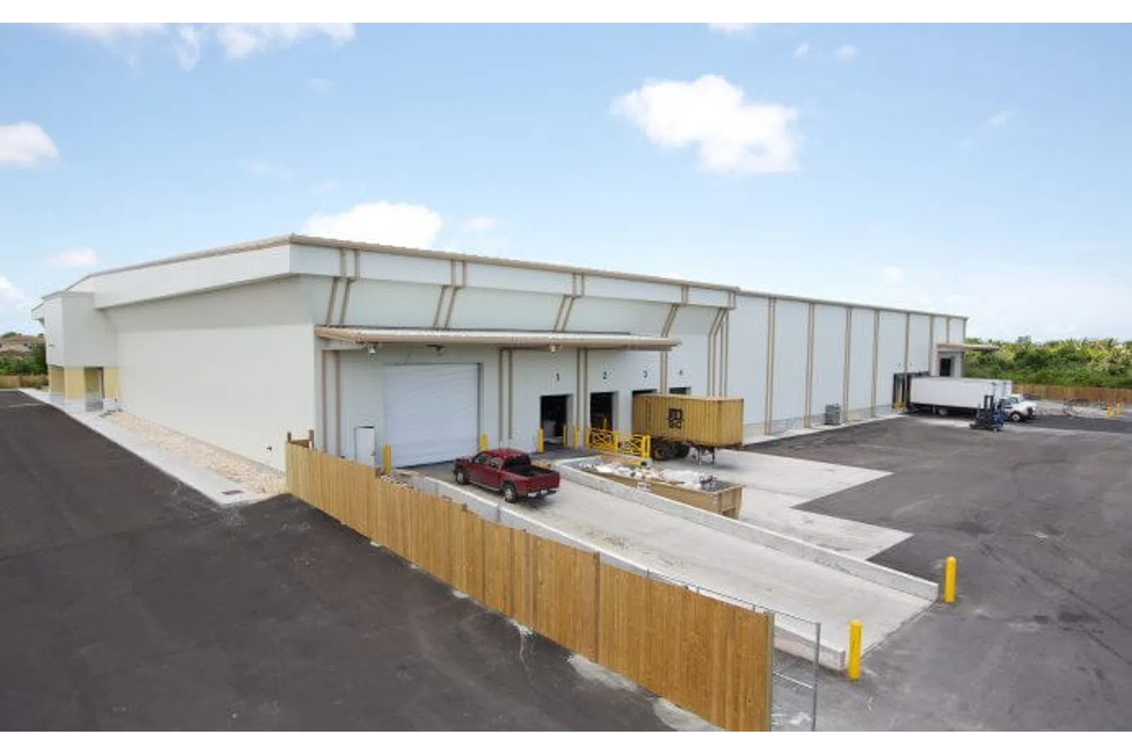 Modern warehouse with loading docks, trucks, and fenced parking area under a blue sky.