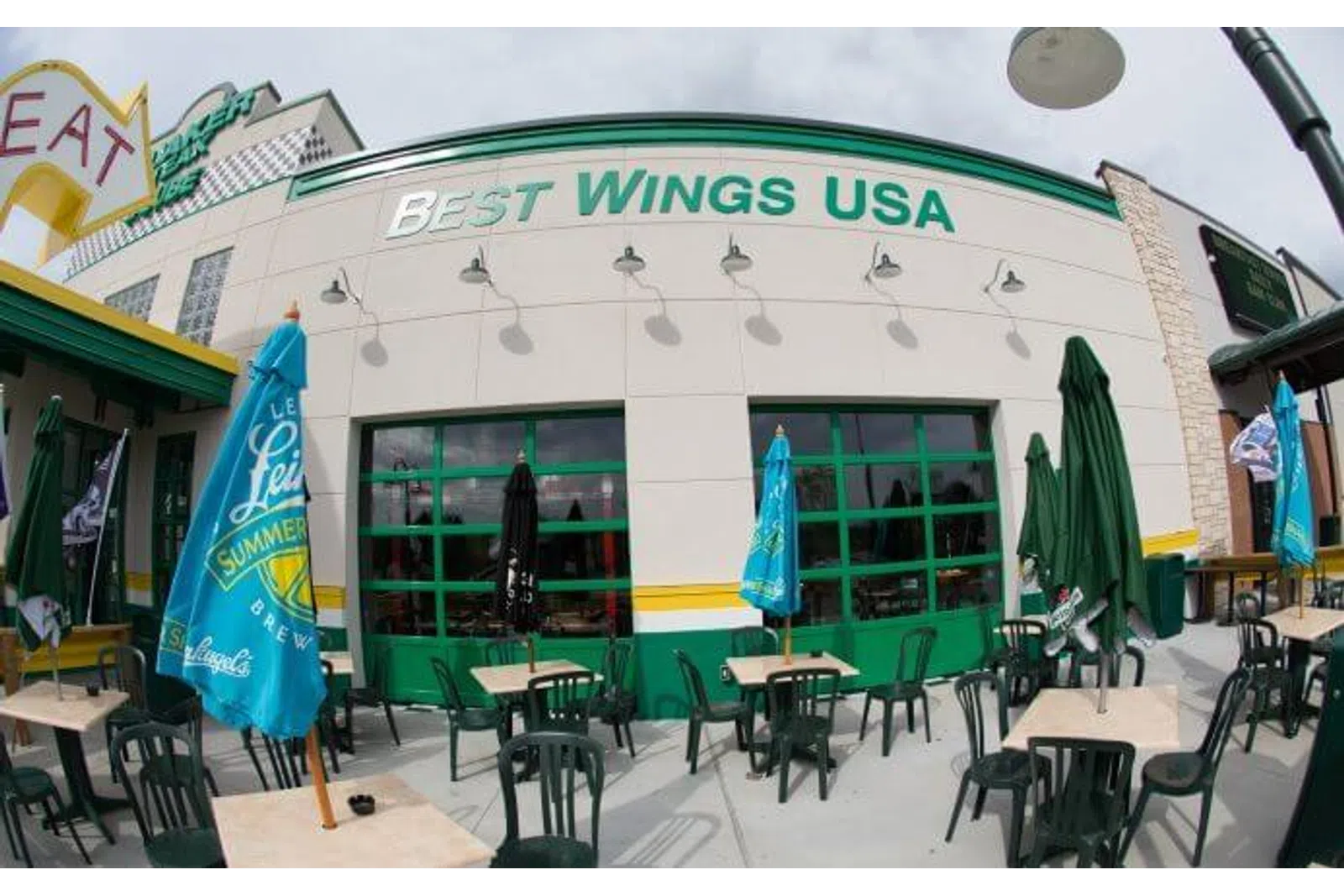 Outdoor seating area at Best Wings USA restaurant with green chairs, tables, and colorful umbrellas on a sunny day.
