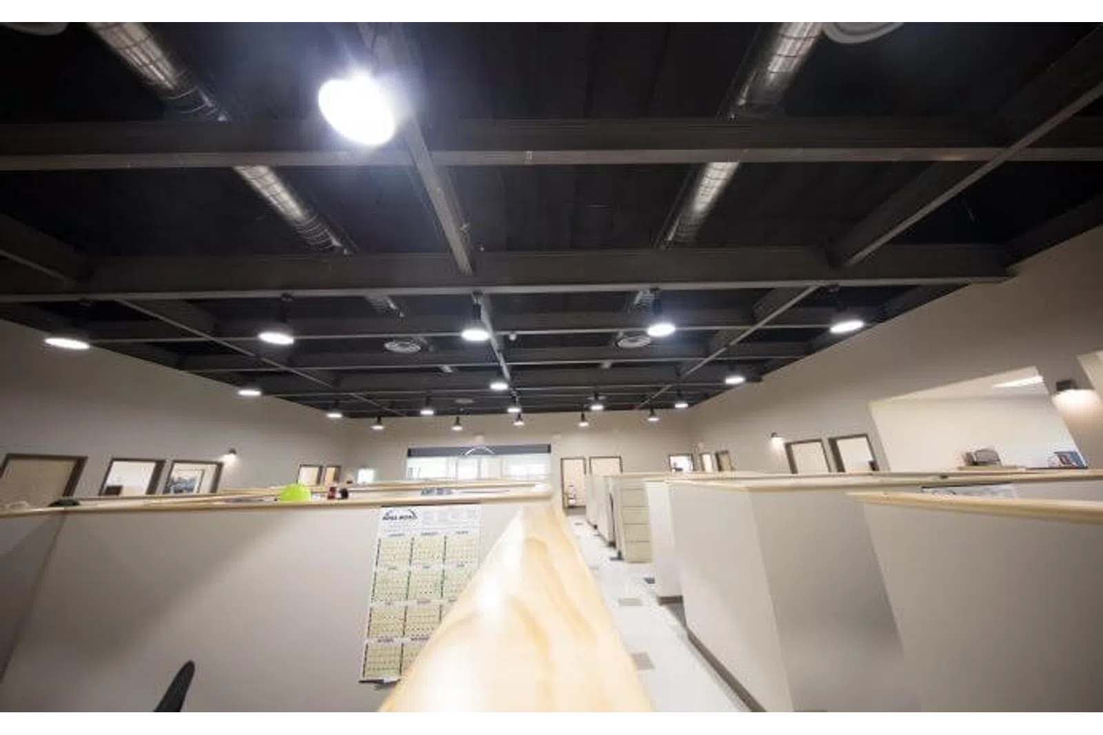 Brightly lit modern office space with multiple cubicles and exposed ceiling ducts under fluorescent lighting.