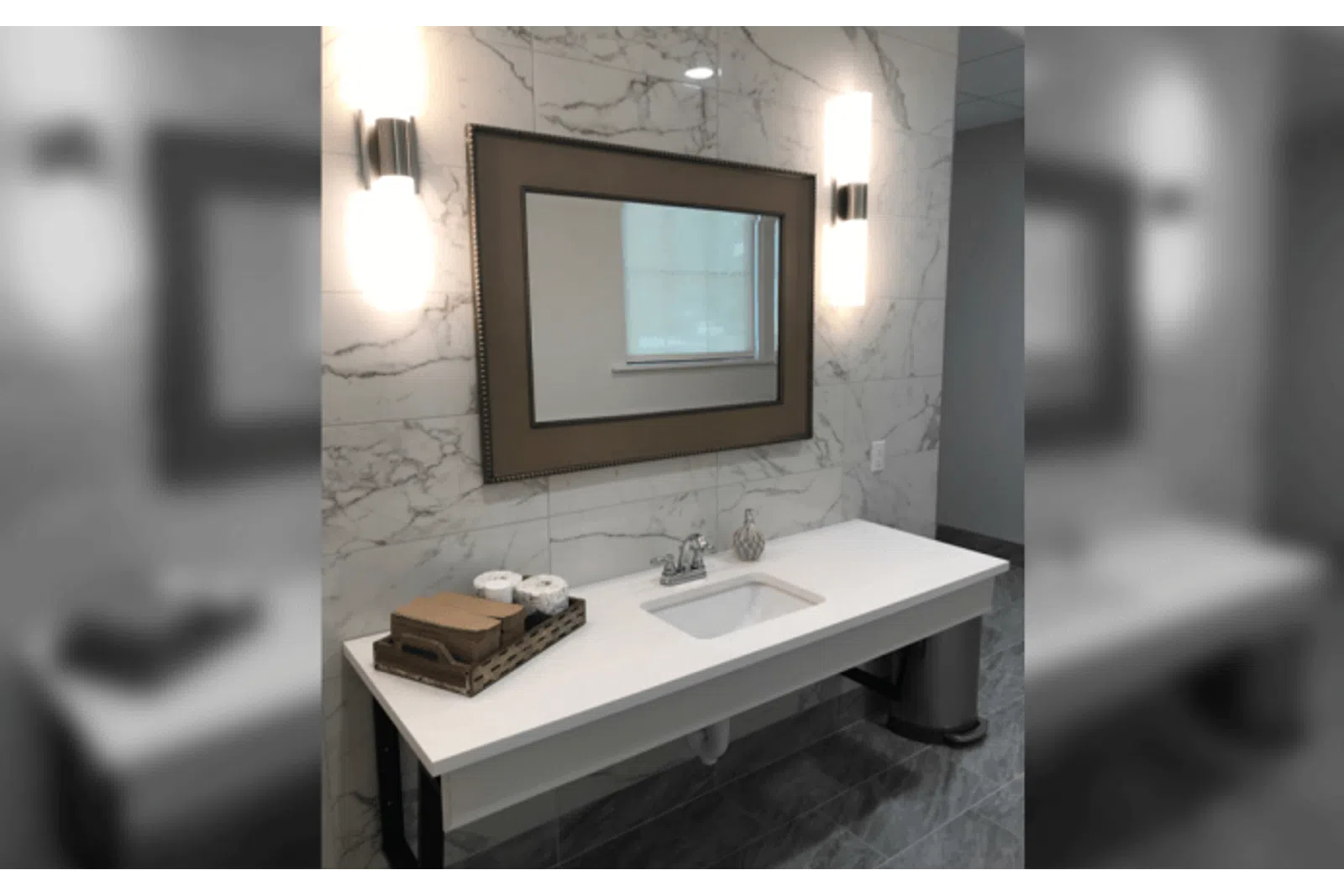 Modern bathroom vanity with white countertop, rectangular sink, framed mirror, and marble wall tiles with sconces.