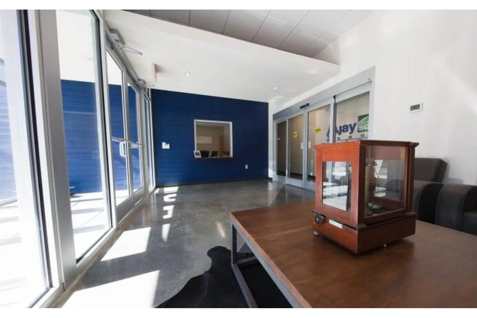 Modern office waiting area with large glass doors, blue accent wall, gray couch, and wooden table with case.
