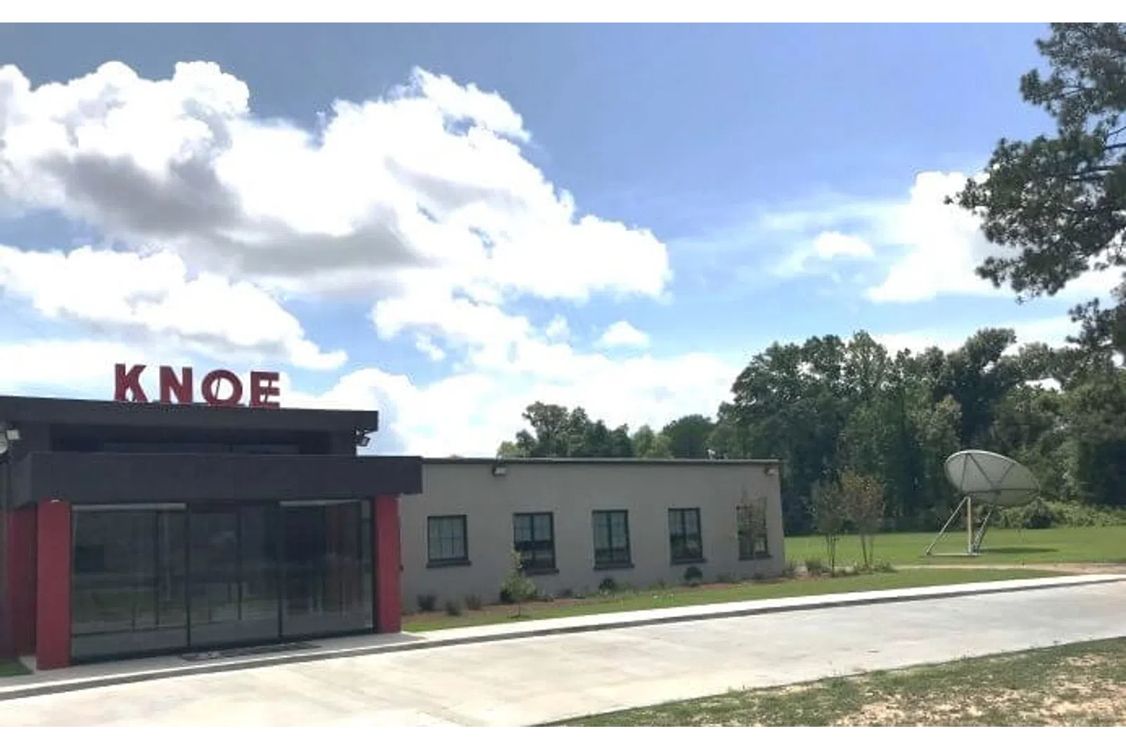 Exterior view of a small building with KNOE sign on the roof under a blue sky with clouds and surrounding greenery.