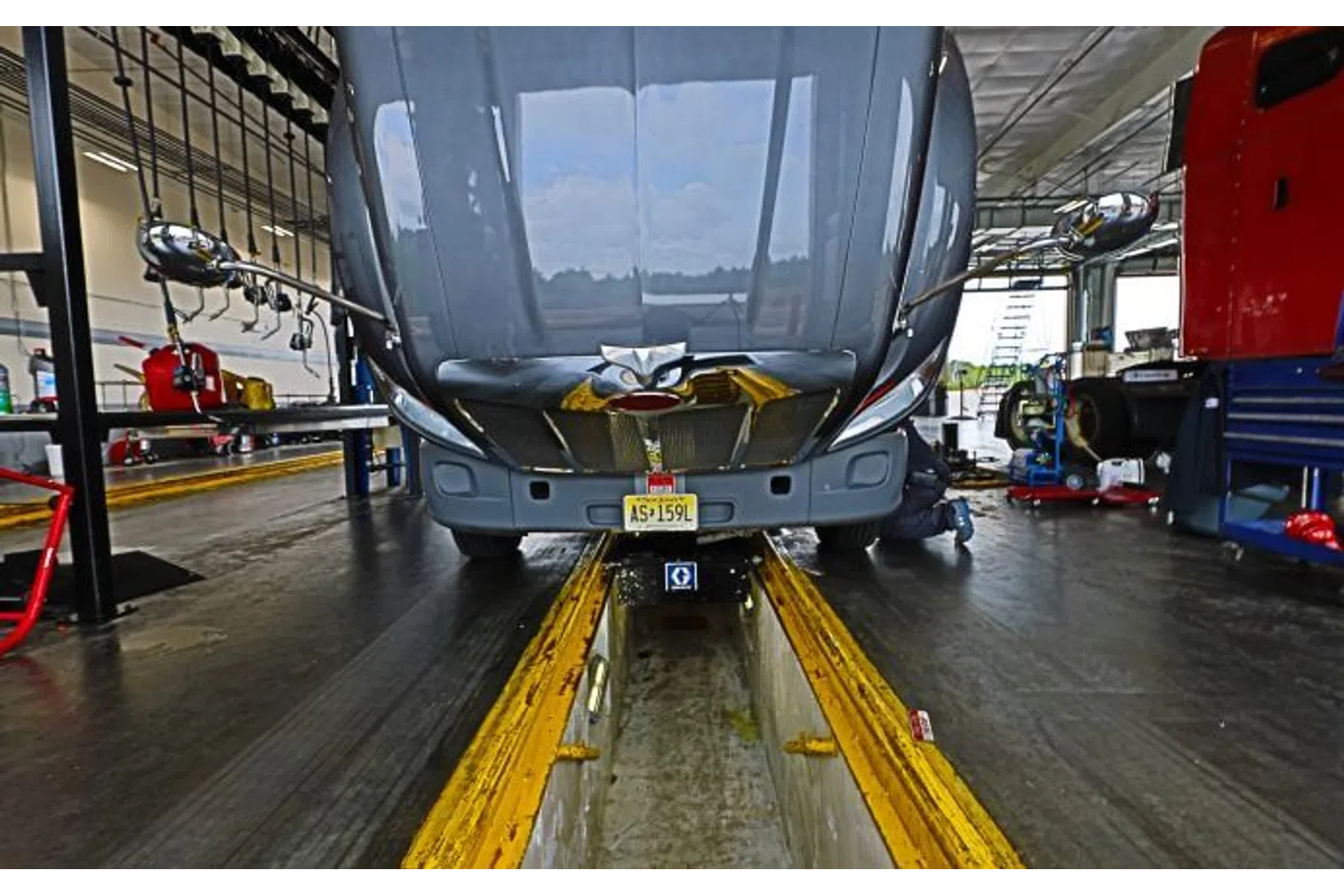 Front view of a truck with lifted hood in a maintenance garage over a service pit with mechanic working underneath.