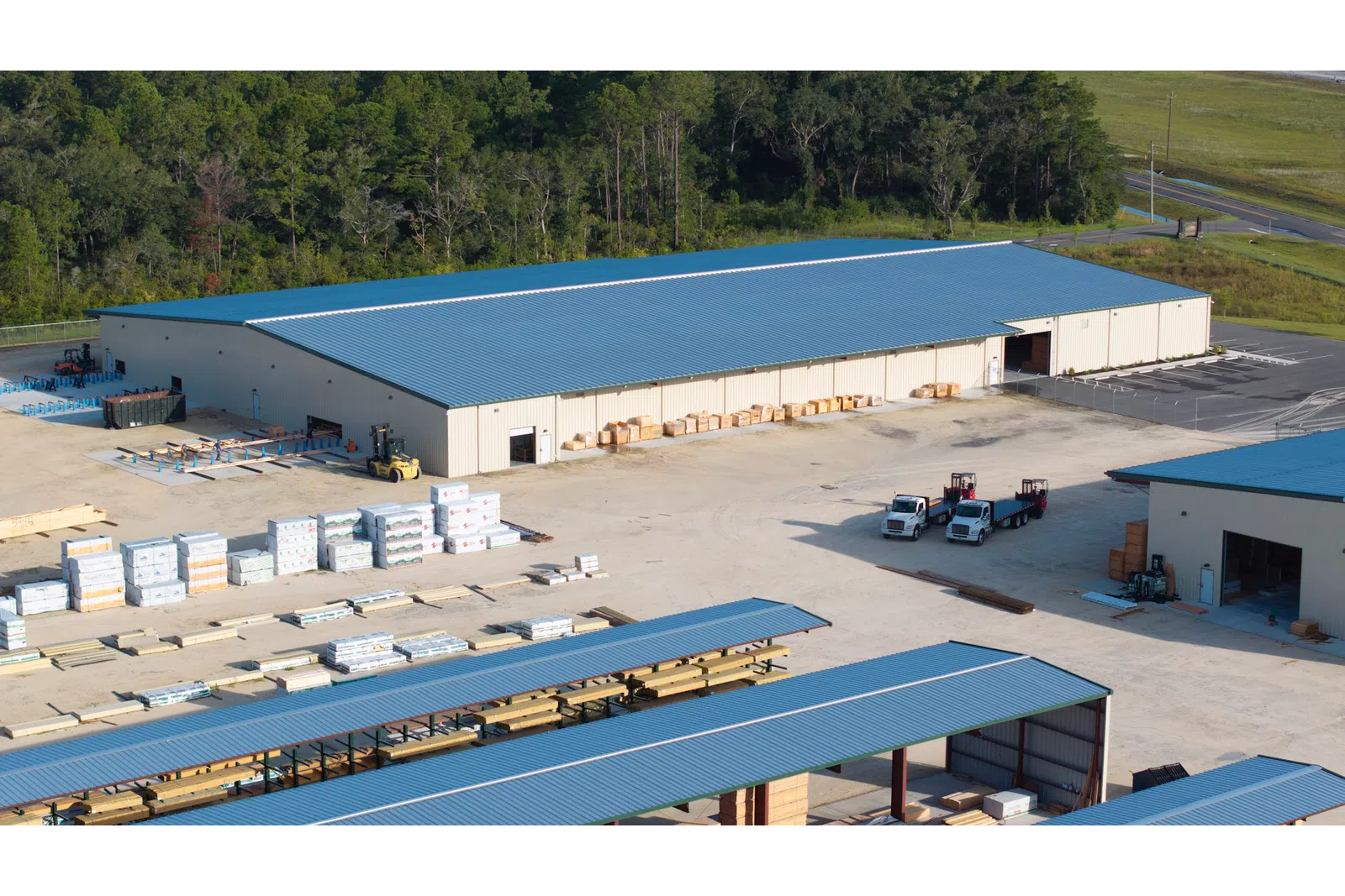 Aerial view of a lumberyard with stacked wood, large storage buildings, and trucks under blue roofs.