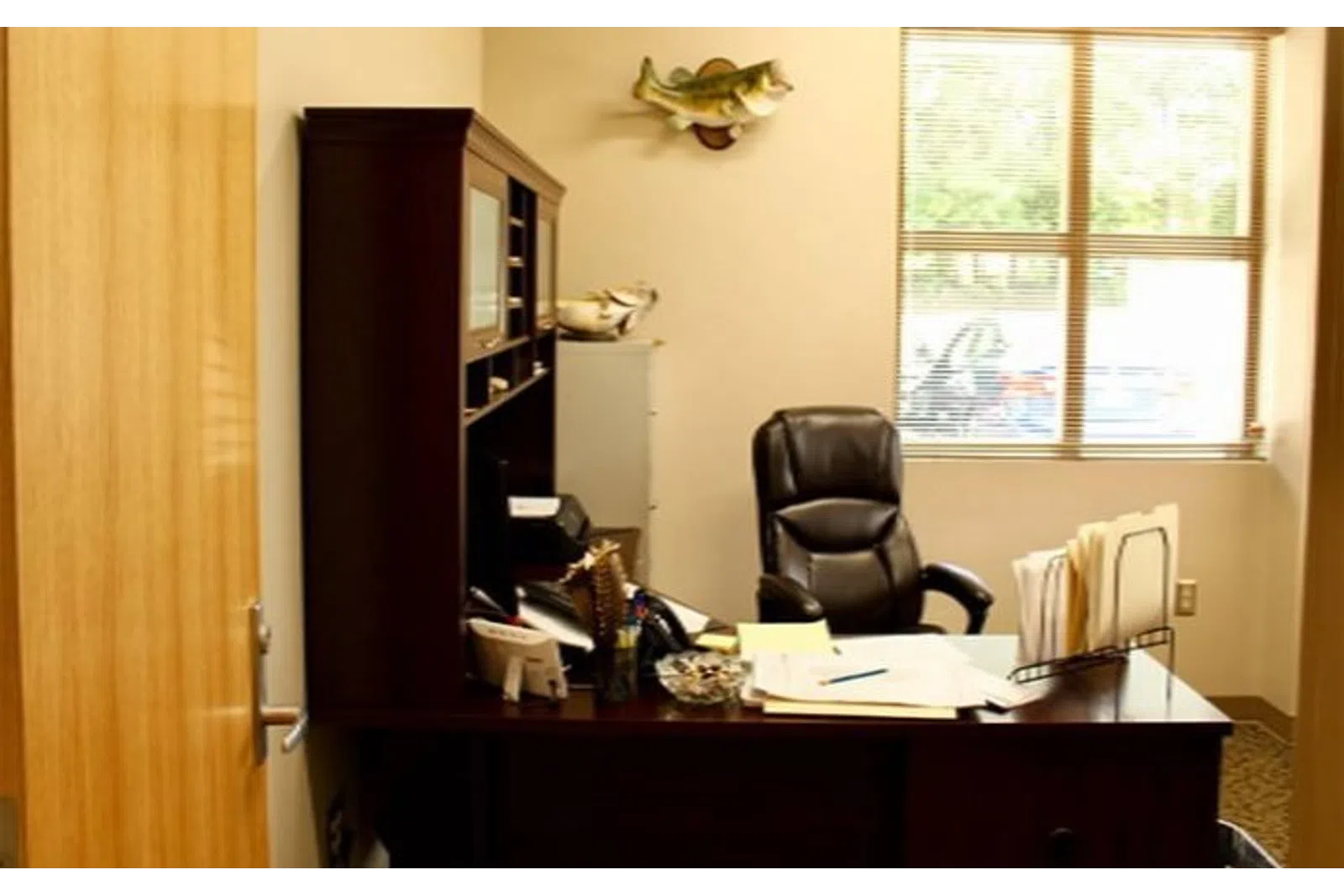 Cozy office setup with dark wood desk, leather chair, paperwork, filing system, and a fish mounted on the wall.