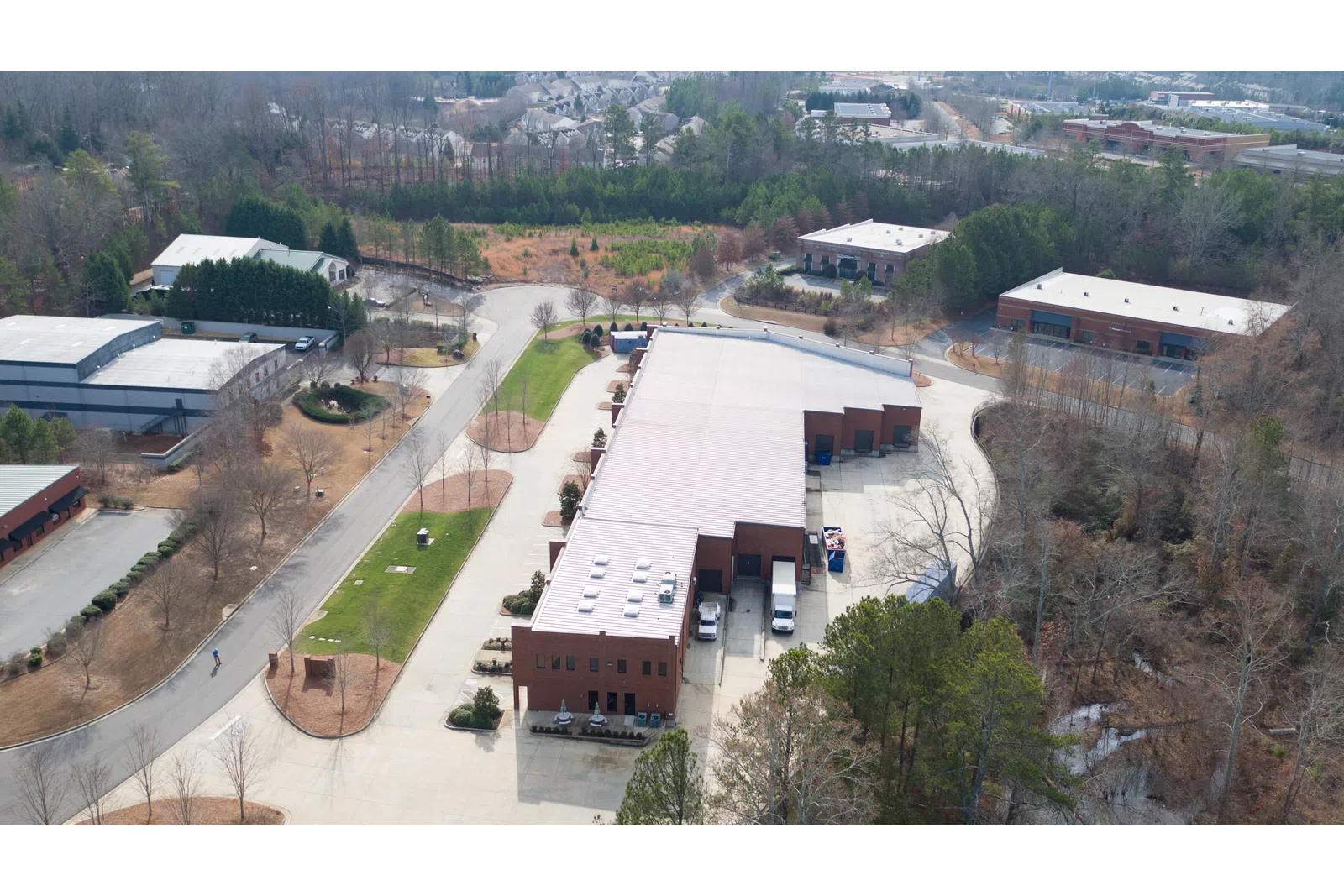 Aerial view of a large industrial warehouse complex surrounded by trees and nearby residential area under clear sky.